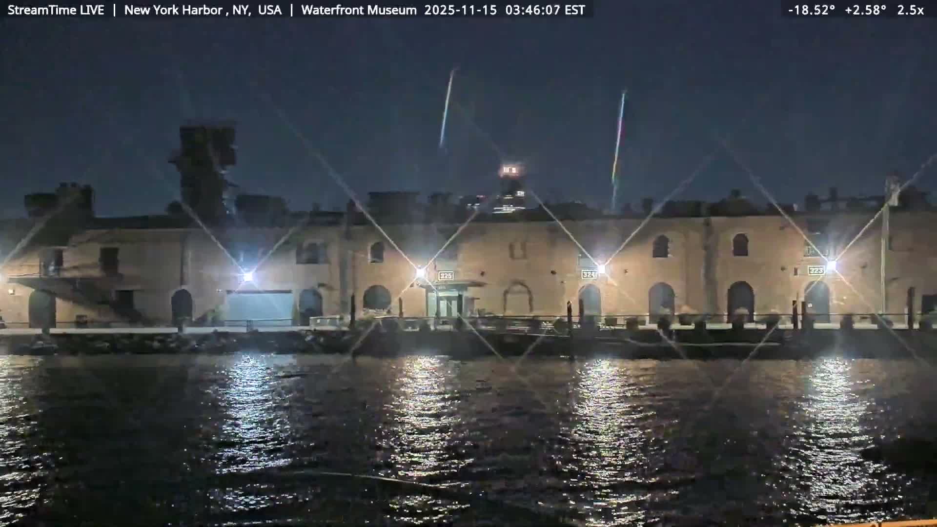 Brightly illuminated waterfront buildings cast shimmering reflections onto dark, rippling water during a rainy night, with visible streaks of precipitation in the air against the dark sky.