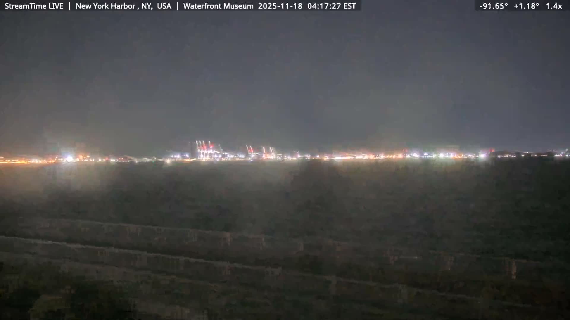 Brightly illuminated waterfront buildings cast shimmering reflections onto dark, rippling water during a rainy night, with visible streaks of precipitation in the air against the dark sky.
