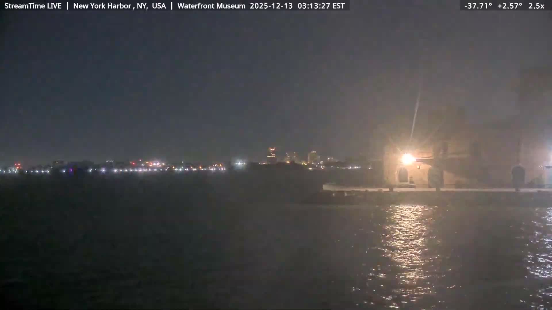 A dark, misty night scene over a harbor features distant city lights and a brightly illuminated structure on the right, with reflections shimmering on the water.