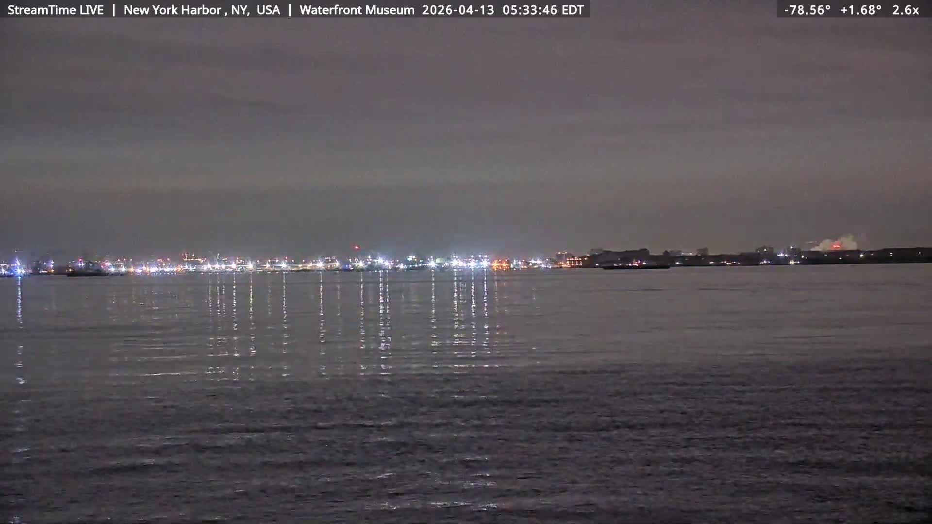 A dark, misty night scene over a harbor features distant city lights and a brightly illuminated structure on the right, with reflections shimmering on the water.