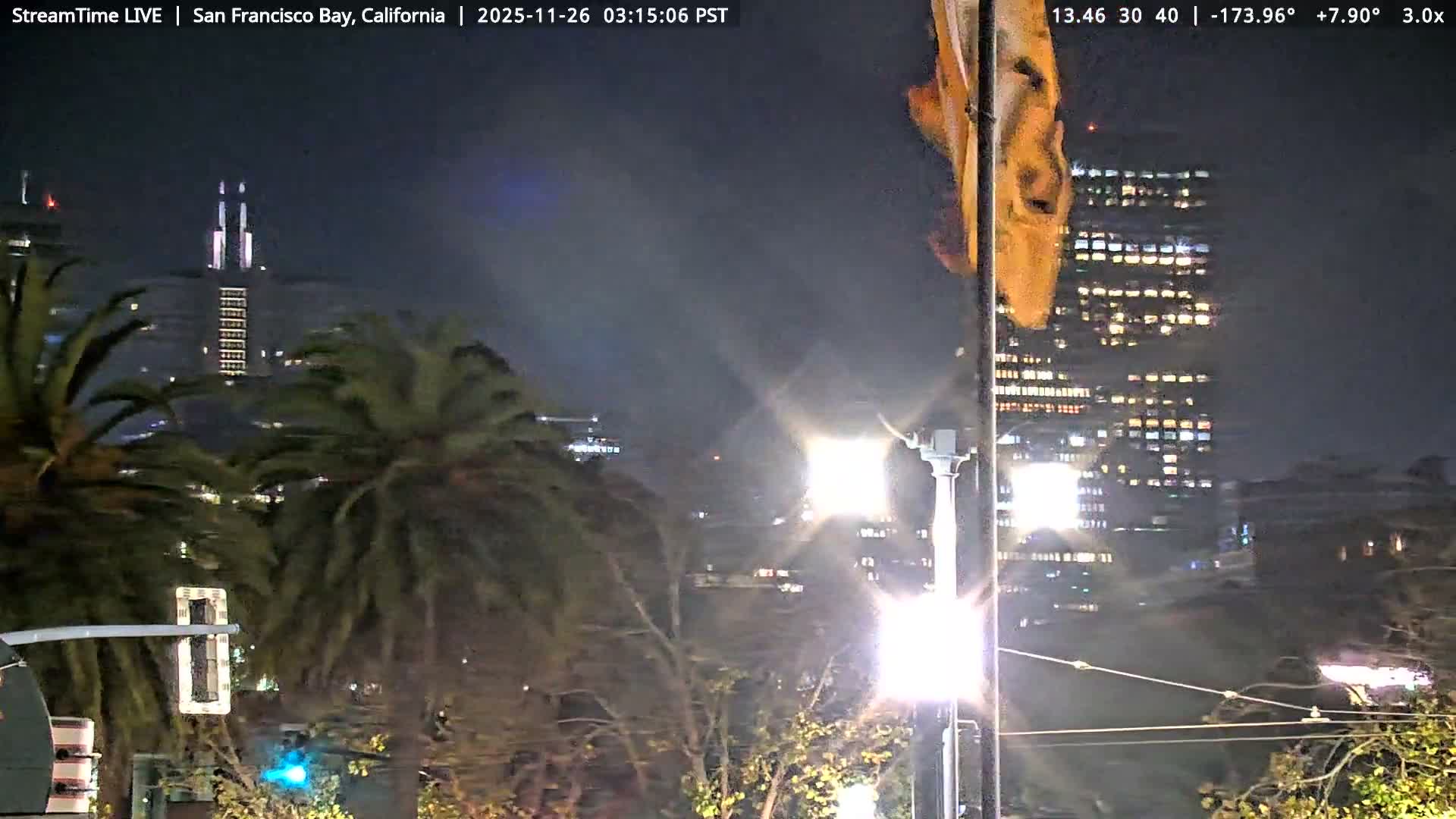 A dark and overcast night view over a body of water shows a distant illuminated city skyline across the bay, with a long pier building visible along the right edge.