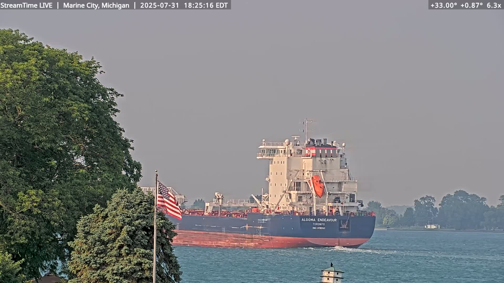 A large cargo ship sails on calm water past a shoreline with green trees and an American flag.