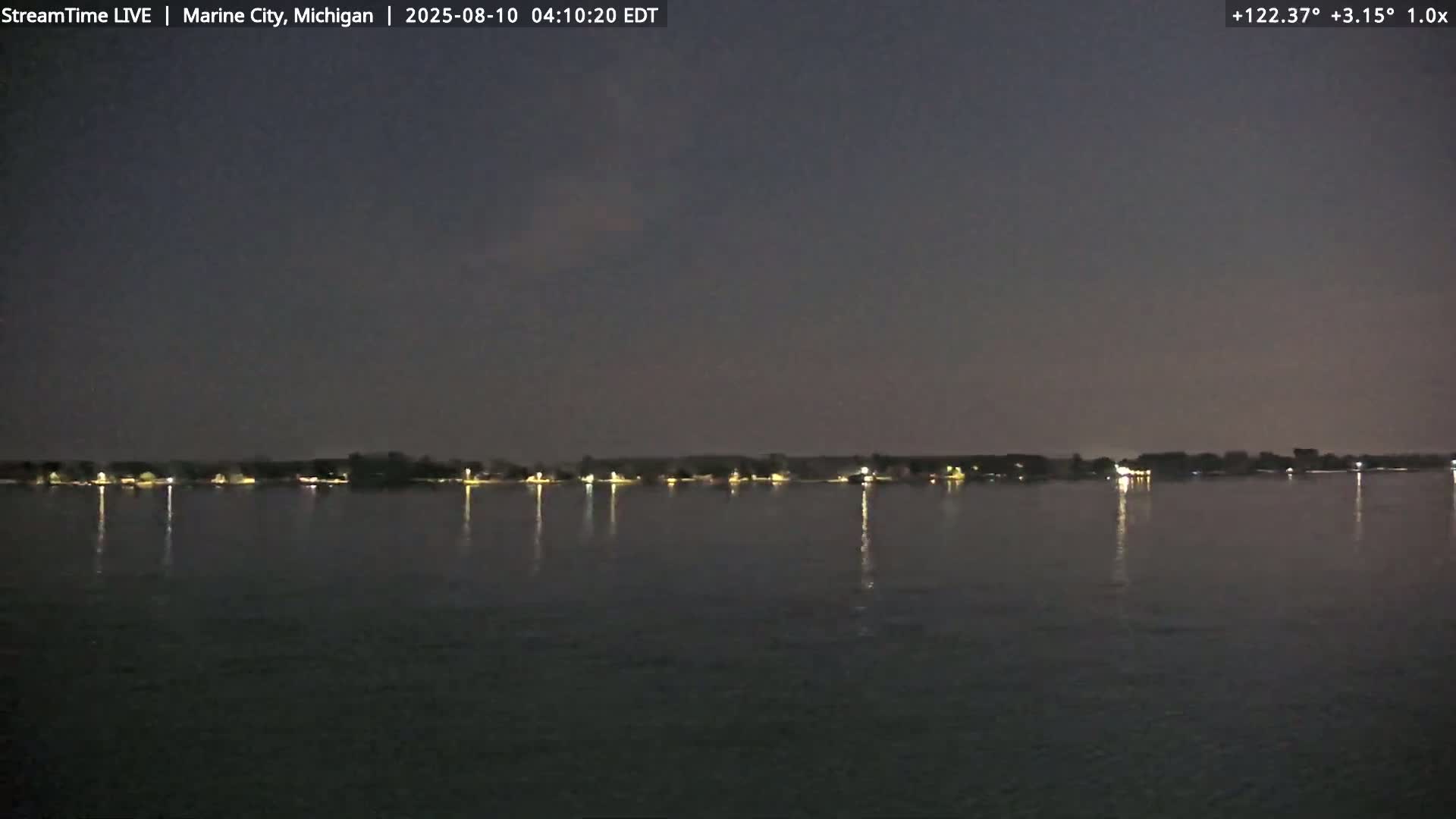 A dark, calm body of water reflects the lights of a distant shoreline at night.