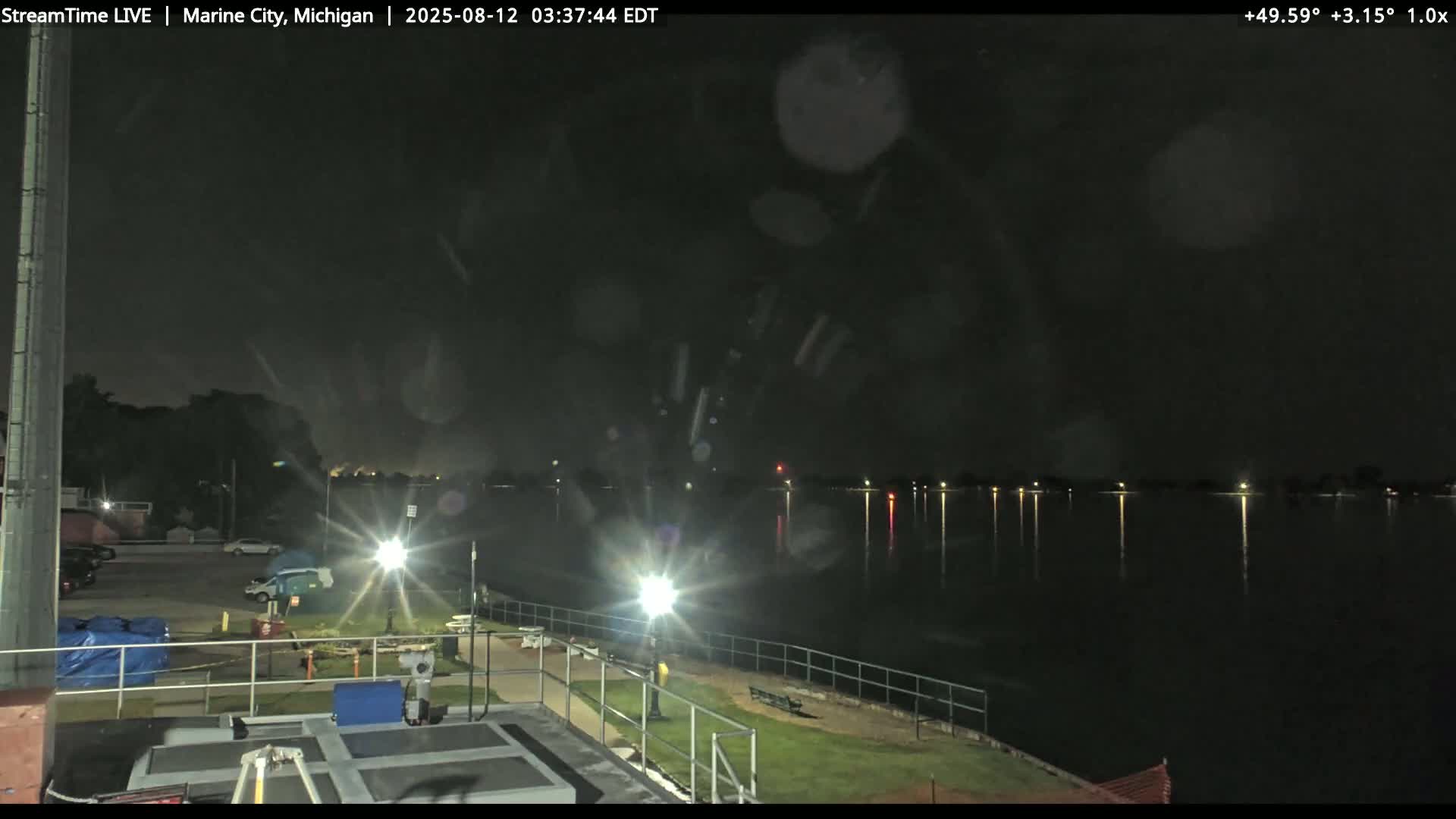 A nighttime view of a calm body of water under a dark, rainy sky, with lights illuminating a nearby waterfront area.