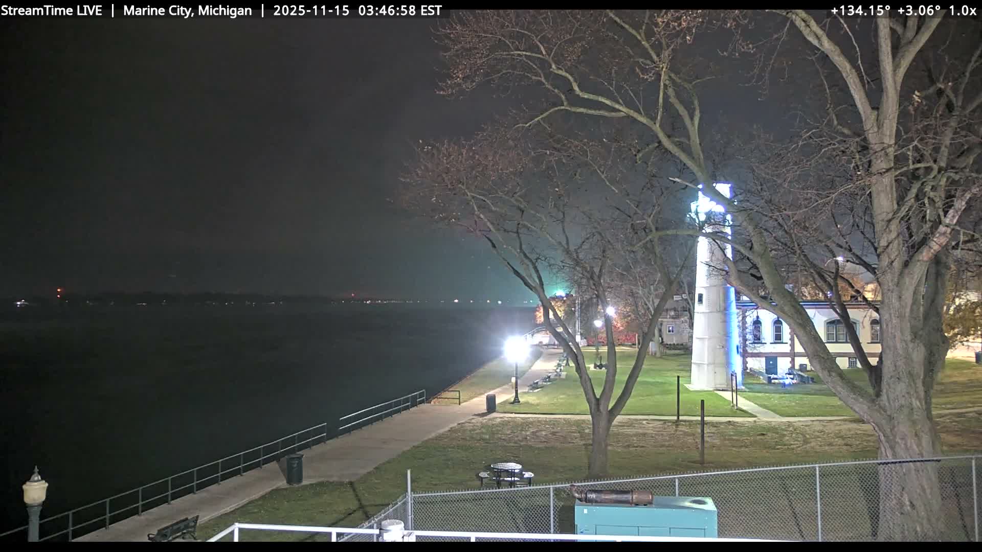 A clear night illuminates a waterfront scene featuring a glowing white lighthouse, bare trees, and a lit promenade alongside dark water with distant shore lights.