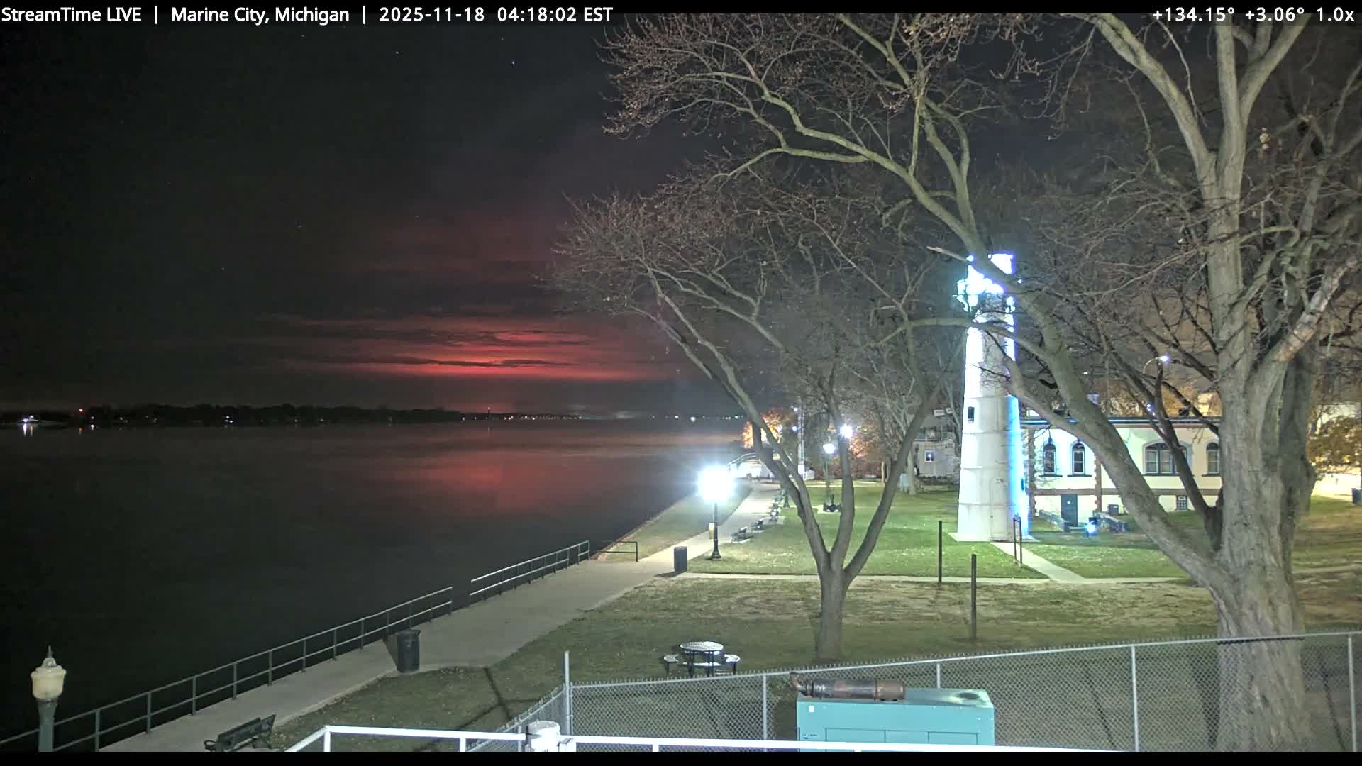 A clear night illuminates a waterfront scene featuring a glowing white lighthouse, bare trees, and a lit promenade alongside dark water with distant shore lights.
