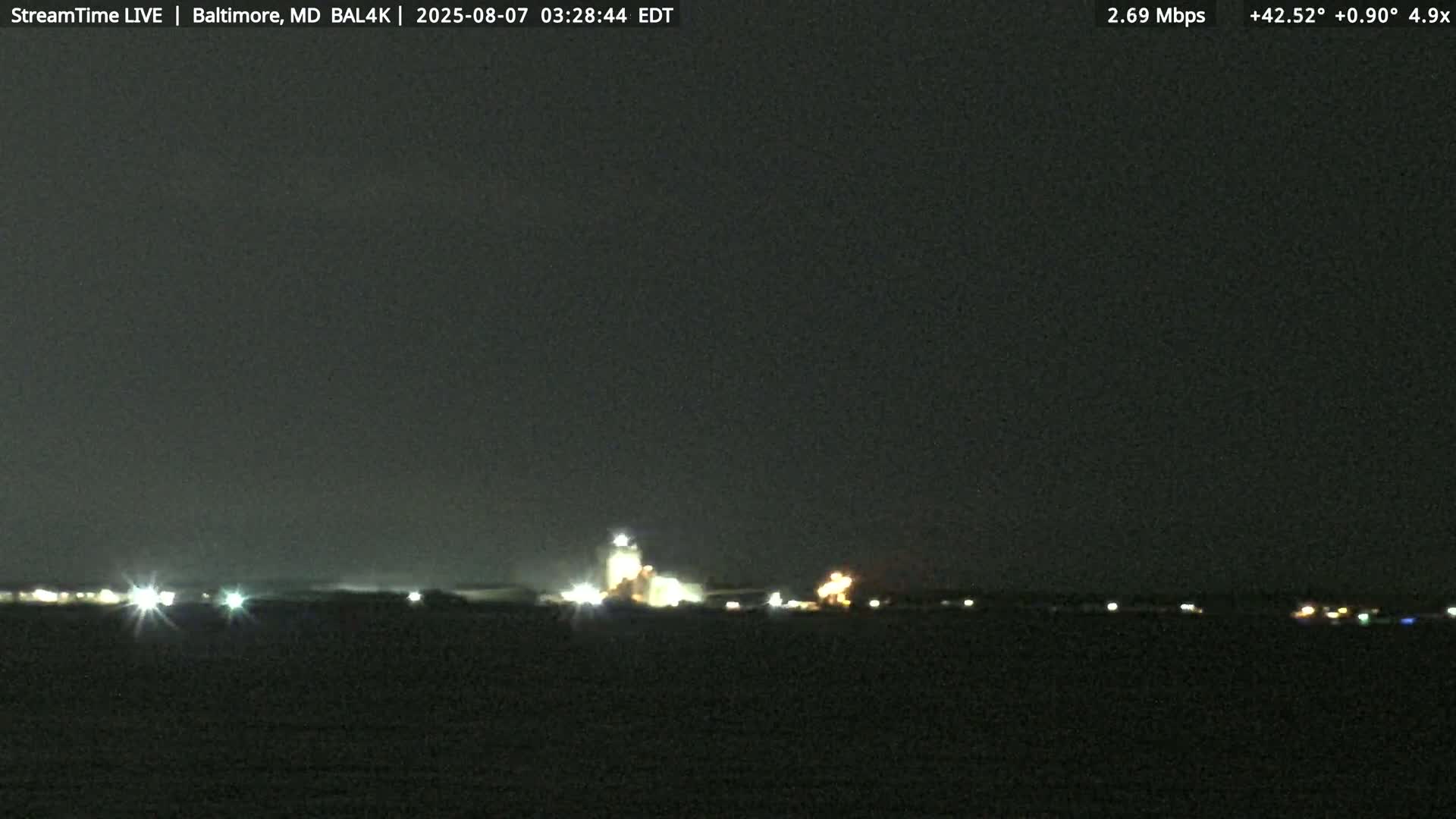 At night, an industrial complex with bright lights is visible across a dark body of water under a clear, dark sky.