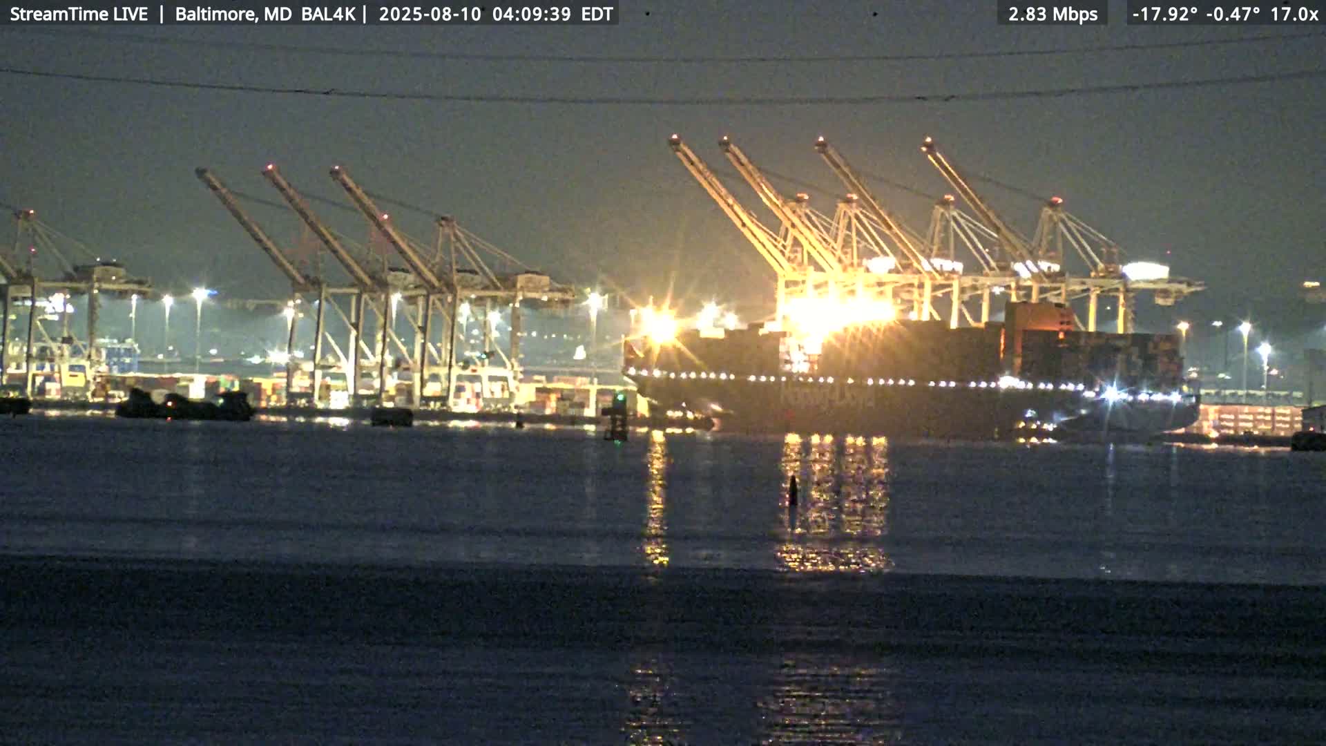 A large container ship is docked at a brightly lit port at night under calm, dark skies.