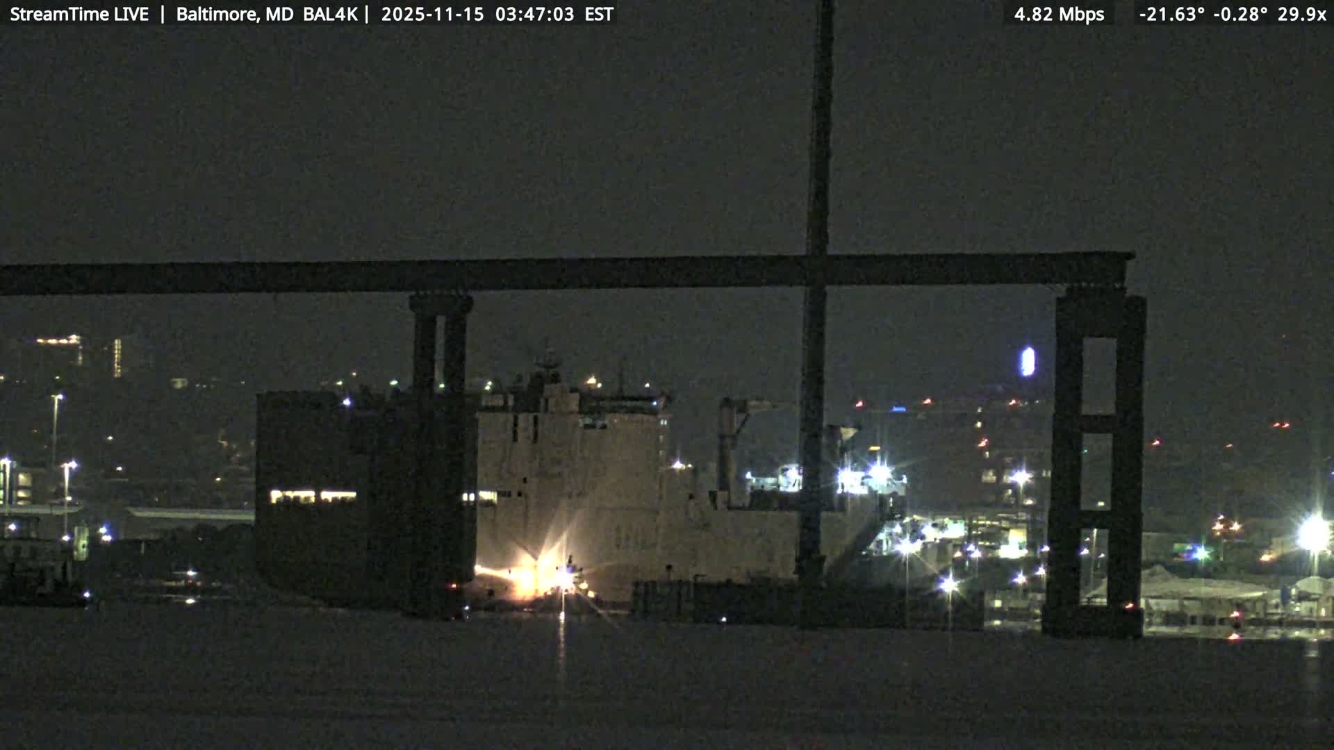A large ship, brightly illuminated by work lights, is visible at night in a dark harbor, partially framed by a large overhead structure, with numerous city or port lights twinkling in the background under clear skies.