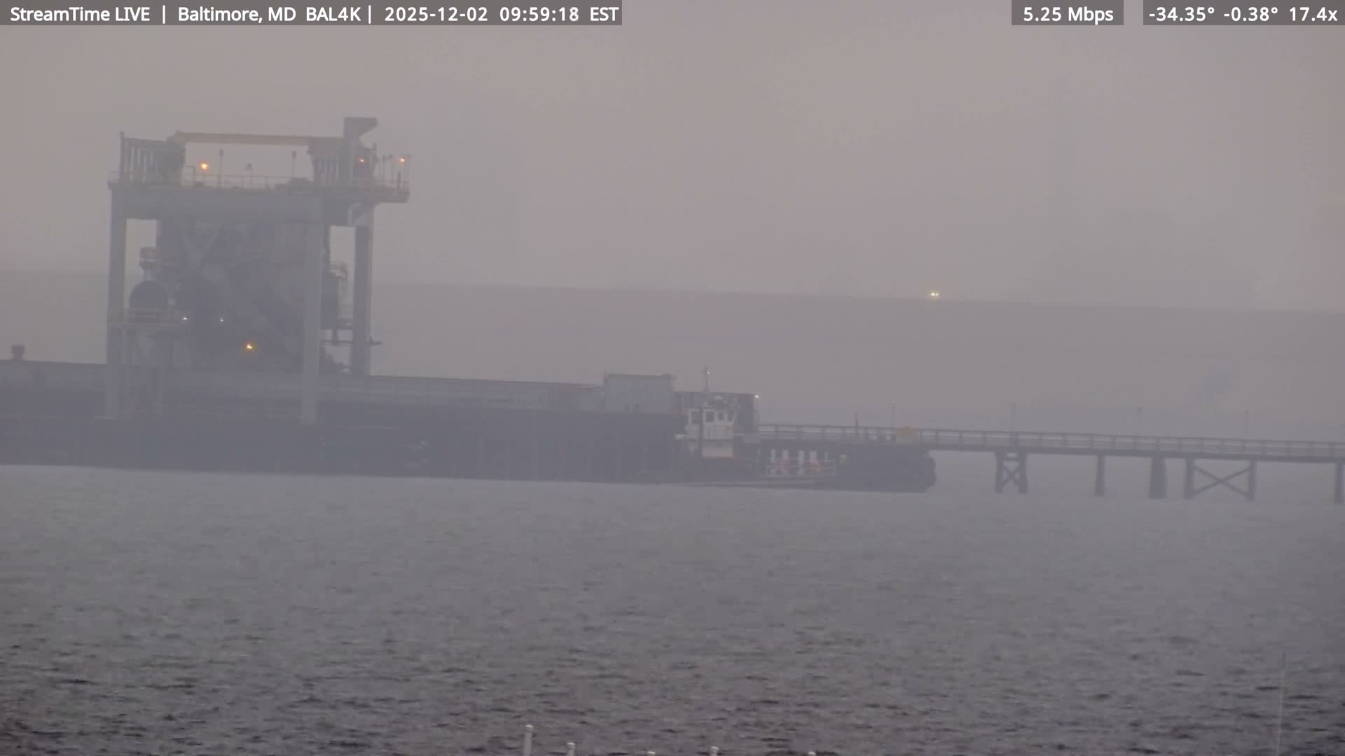 On a very foggy day, a large industrial loading facility with lights illuminated stands next to a docked ship and an extending pier over dark water, all shrouded in heavy mist.