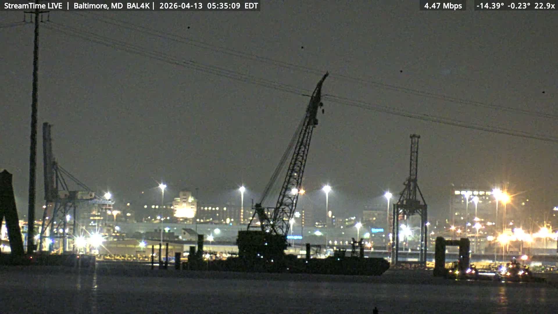 A vast expanse of calm dark water reflects the numerous brightly lit cranes and industrial structures of a busy port under a clear night sky.