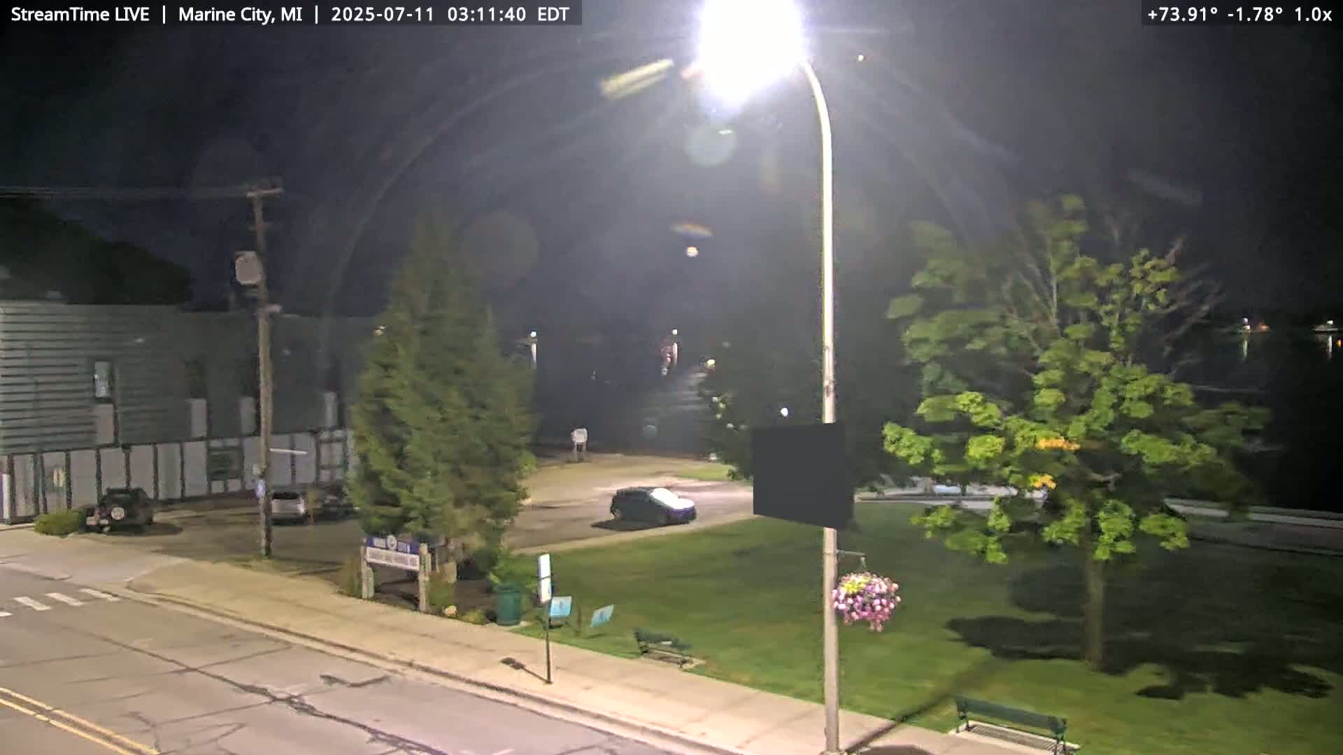A nighttime view of a street scene showing a car in a small park next to a road and a building with some cars in a parking lot.