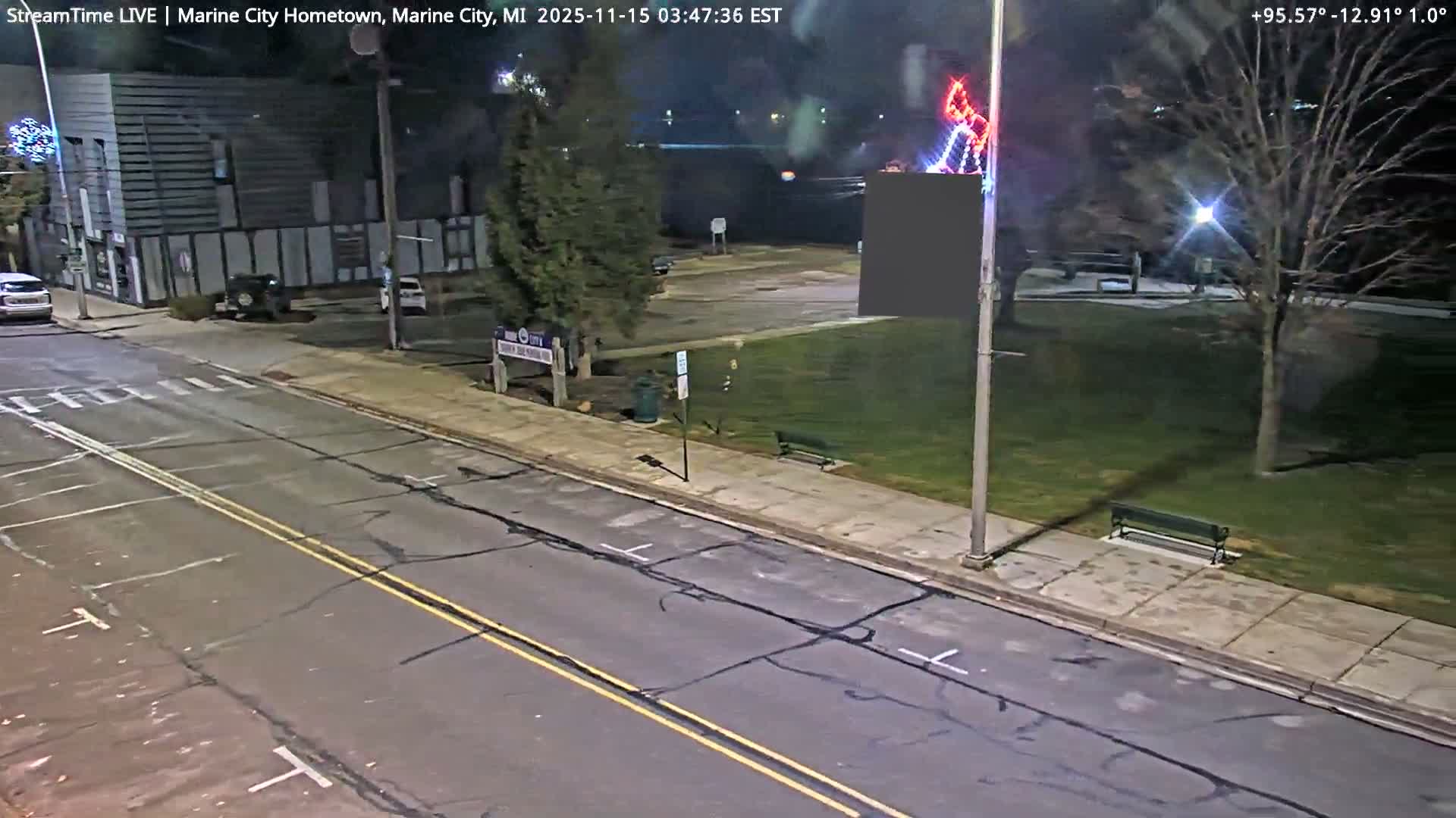 A nocturnal, high-angle view reveals a damp street with cracked asphalt and a few parked cars, flanked by buildings on the left and a park with bare trees, benches, and colorful decorative lights on the right, all under a clear, dark sky.