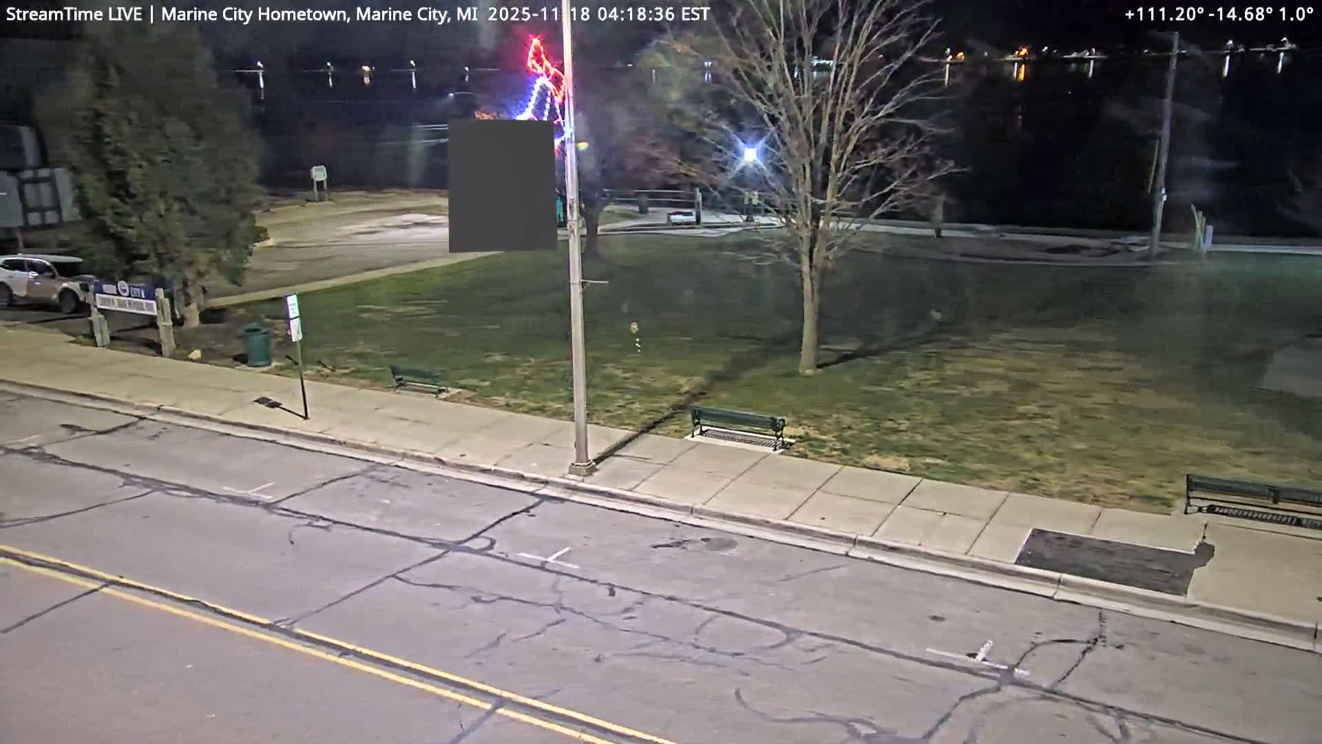 A nocturnal, high-angle view reveals a damp street with cracked asphalt and a few parked cars, flanked by buildings on the left and a park with bare trees, benches, and colorful decorative lights on the right, all under a clear, dark sky.