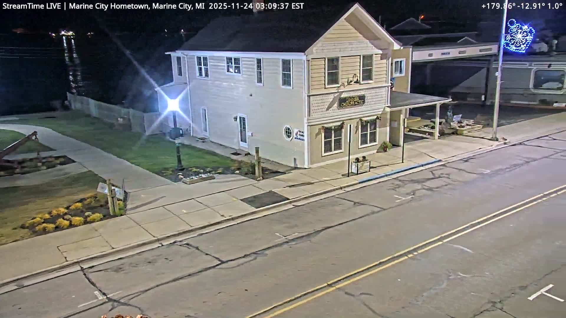 An illuminated two-story building stands alongside a street and sidewalk at night, with a distant body of water visible on the left, under clear weather.