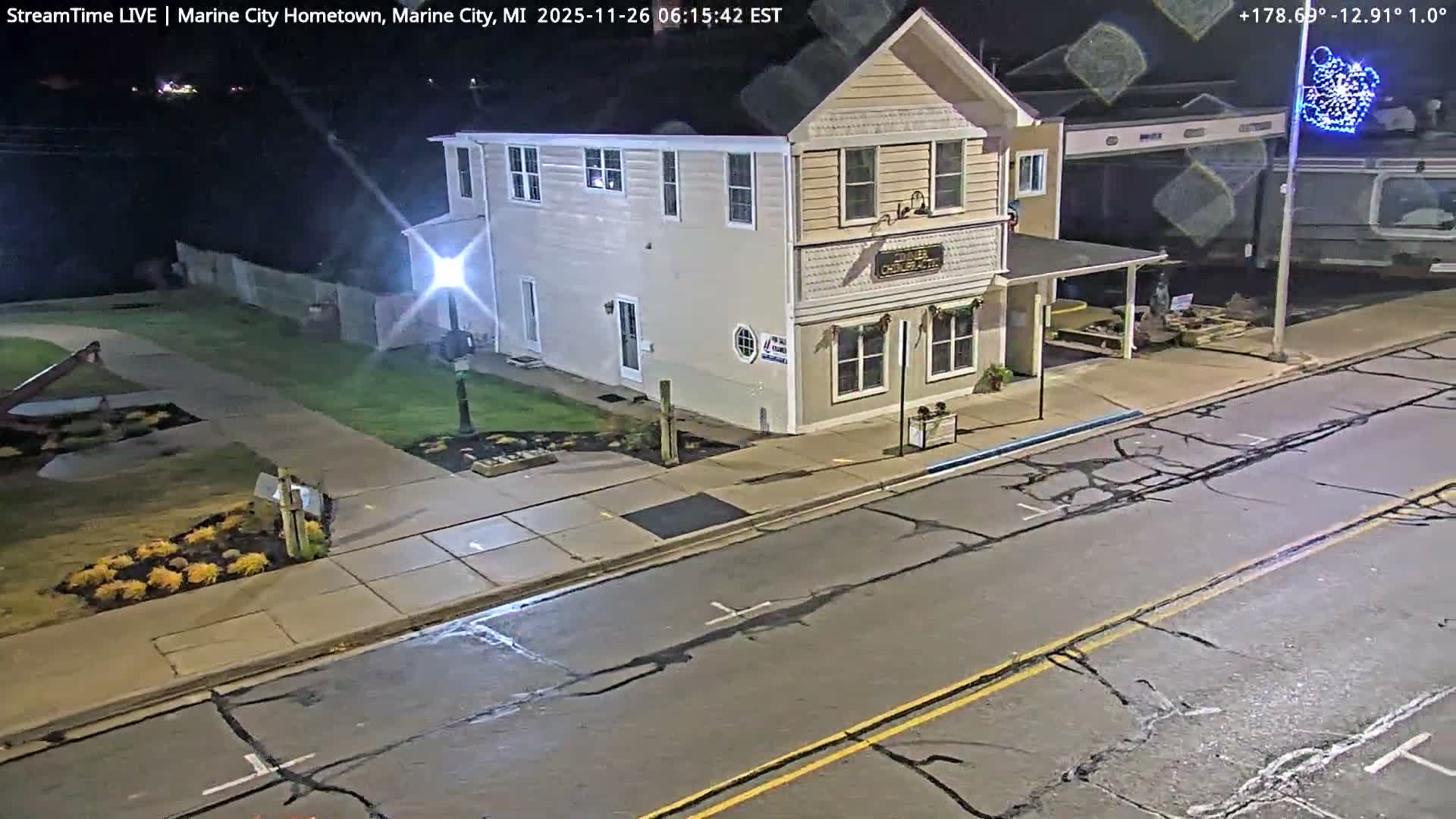 An illuminated two-story building stands alongside a street and sidewalk at night, with a distant body of water visible on the left, under clear weather.