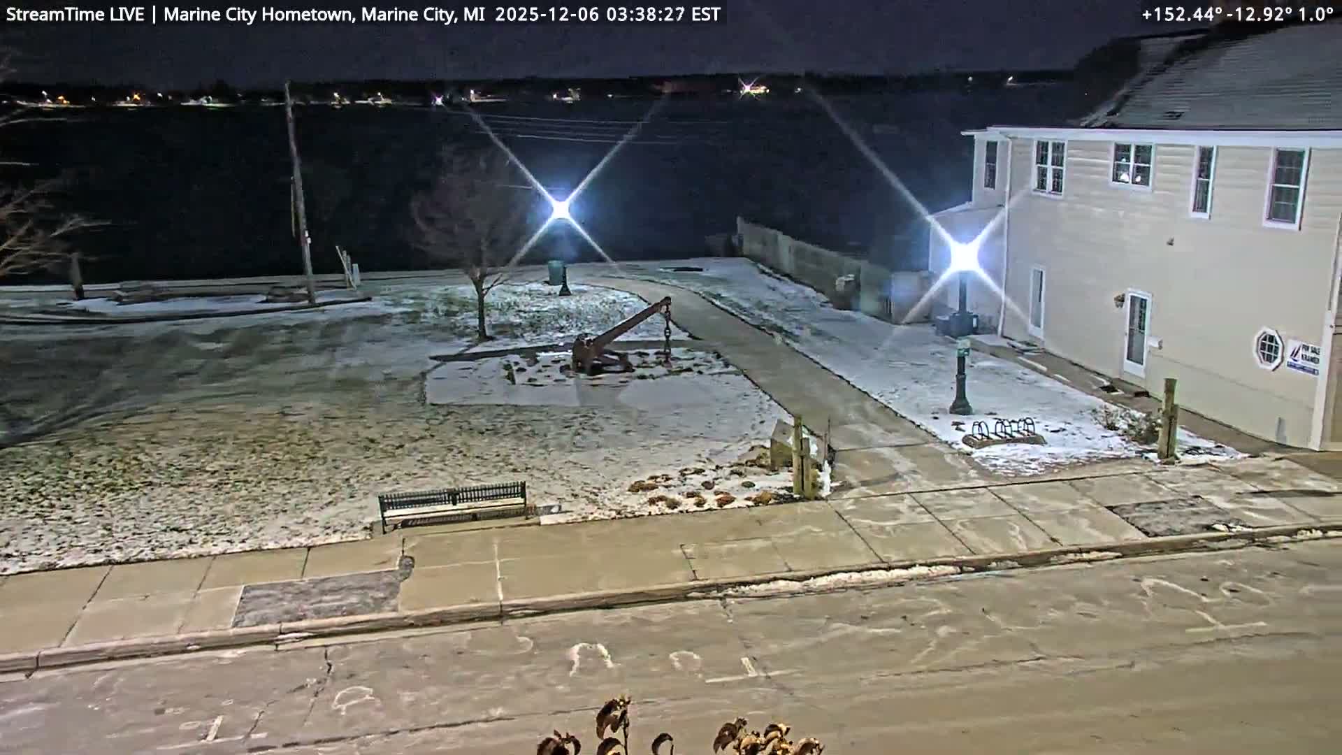 The image displays a lightly snow-dusted park featuring a large anchor sculpture and an adjacent two-story building at night, overlooking a dark body of water with distant shore lights visible.