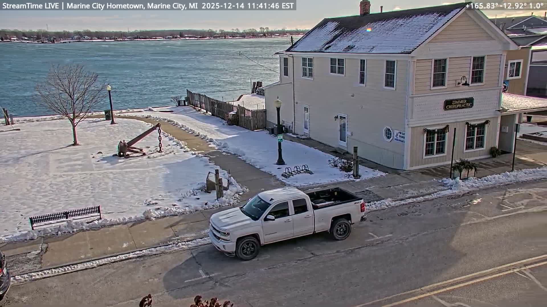 The image displays a lightly snow-dusted park featuring a large anchor sculpture and an adjacent two-story building at night, overlooking a dark body of water with distant shore lights visible.