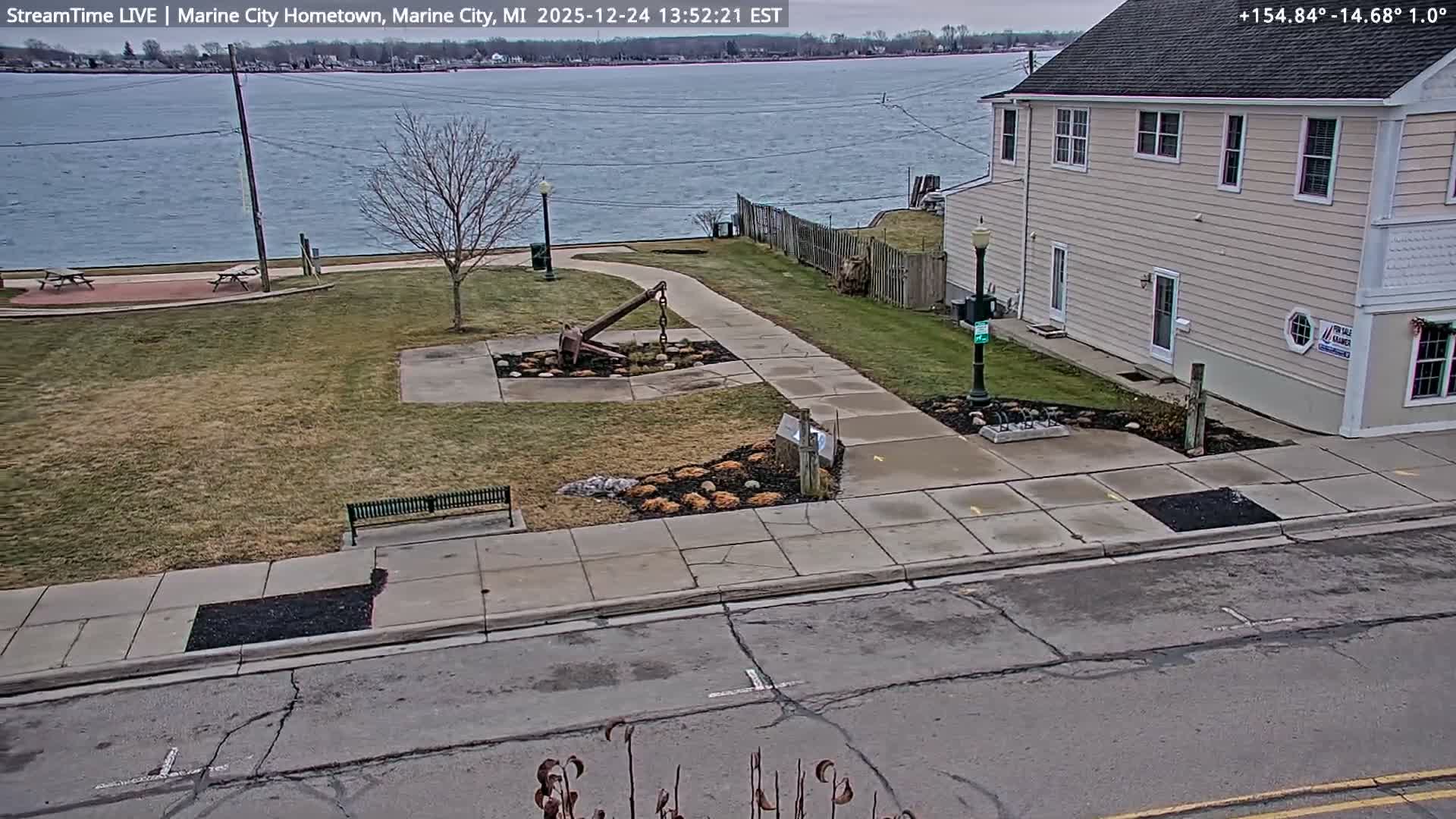 The image displays a lightly snow-dusted park featuring a large anchor sculpture and an adjacent two-story building at night, overlooking a dark body of water with distant shore lights visible.