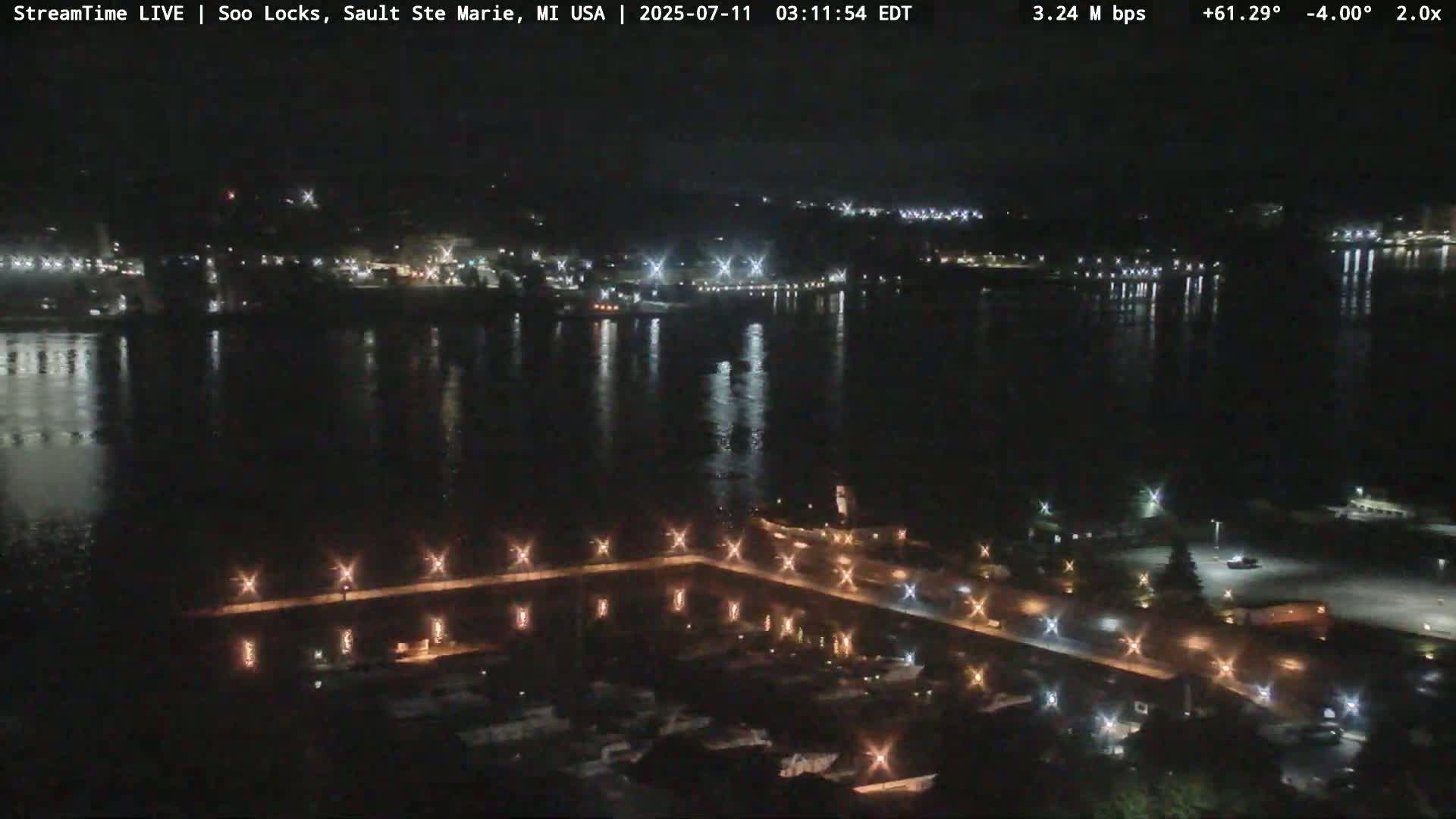 A nighttime view of a dark, calm body of water reflecting city lights across from a landmass with illuminated walkways and buildings.