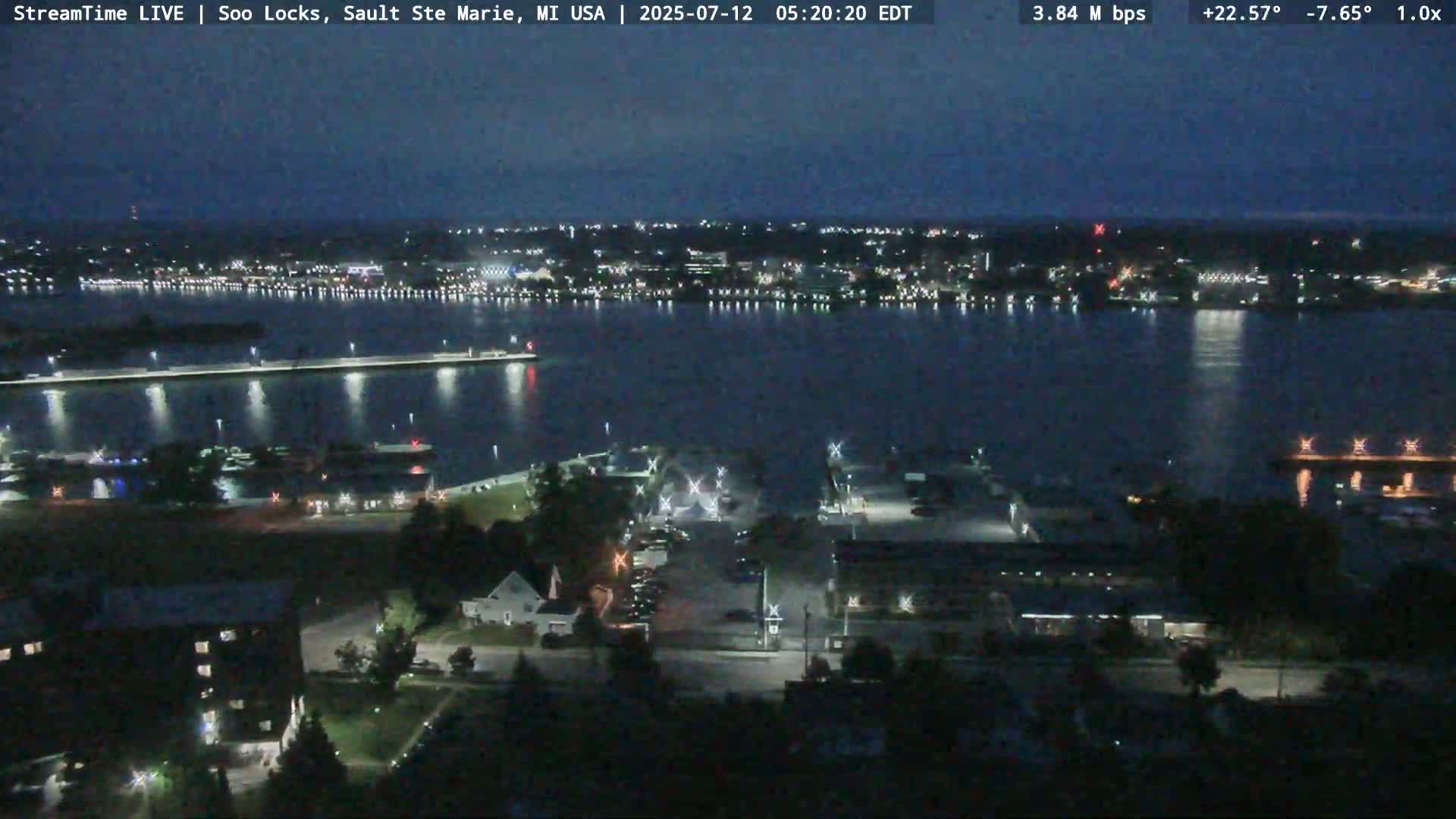 A nighttime view of a dark, calm body of water reflecting city lights across from a landmass with illuminated walkways and buildings.