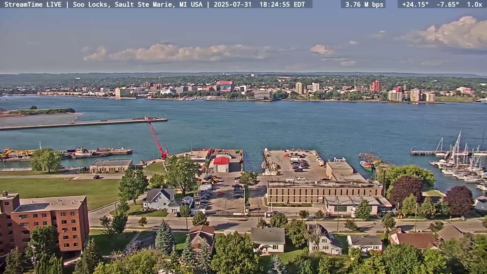 A partly cloudy day shows a waterfront view of a city with a harbor, industrial area, and residential buildings.