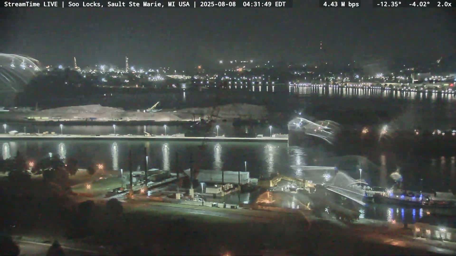 A nighttime, high-angle view shows a waterway with industrial activity and city lights in the background under a clear, dark sky.