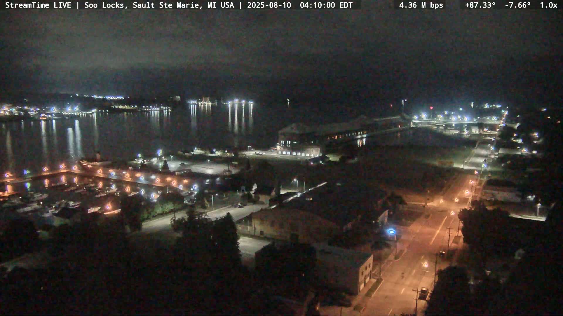 A nighttime aerial view shows a harbor, roads, and buildings near a body of water under a clear, dark sky.