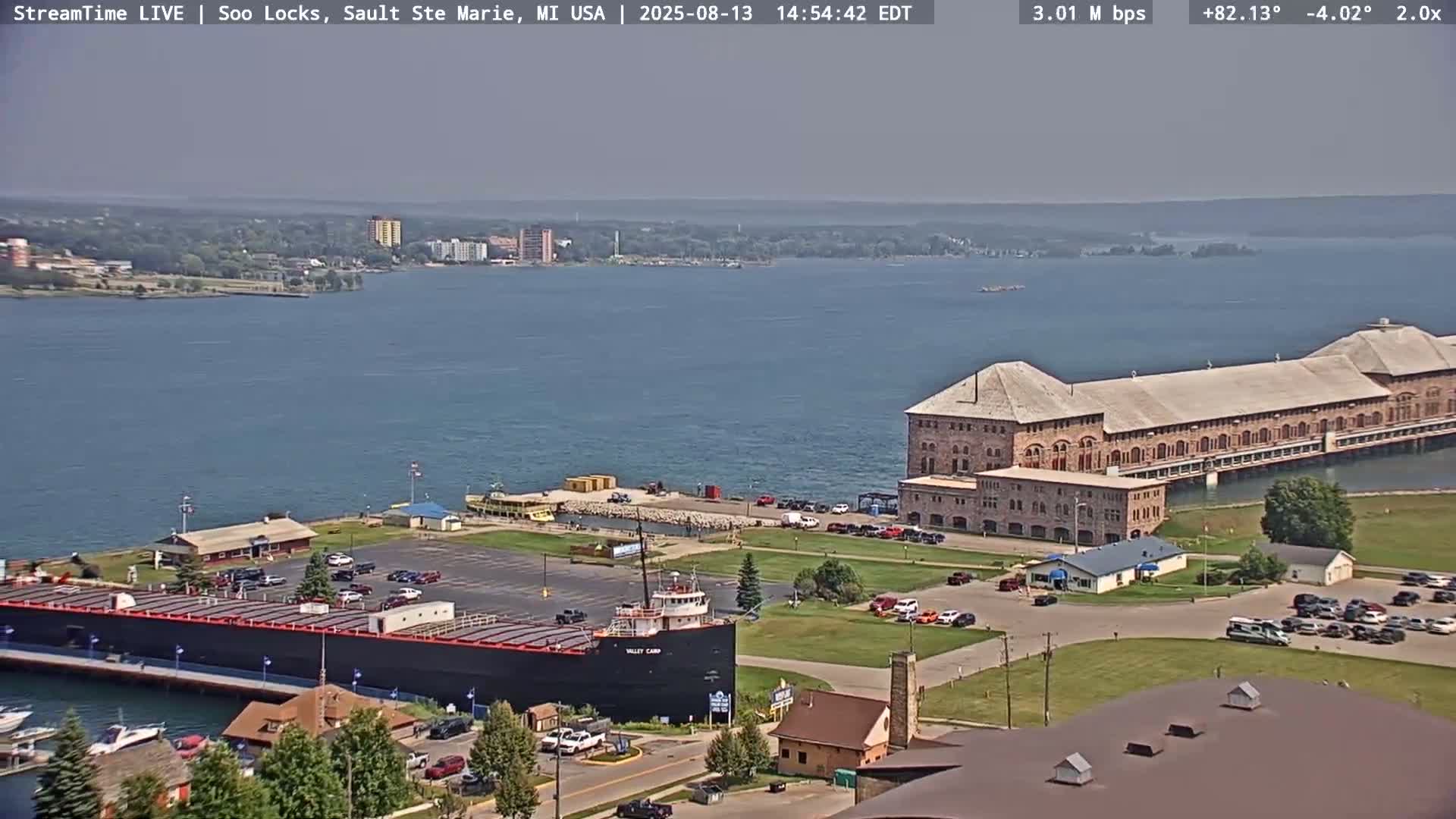 A large body of water is shown with a docked ship, a long brick building, and a cityscape in the distance under hazy, sunny conditions.