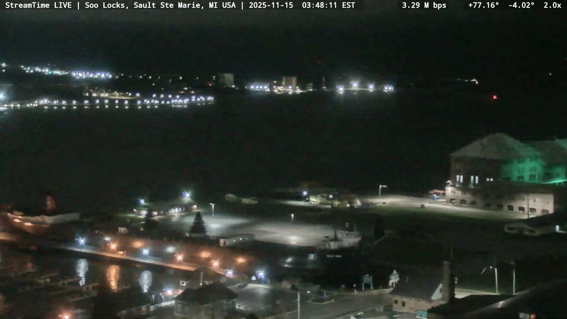 The image shows a clear, dark night view of a waterfront area with numerous lights illuminating buildings, docks with boats, and reflecting across the still water.