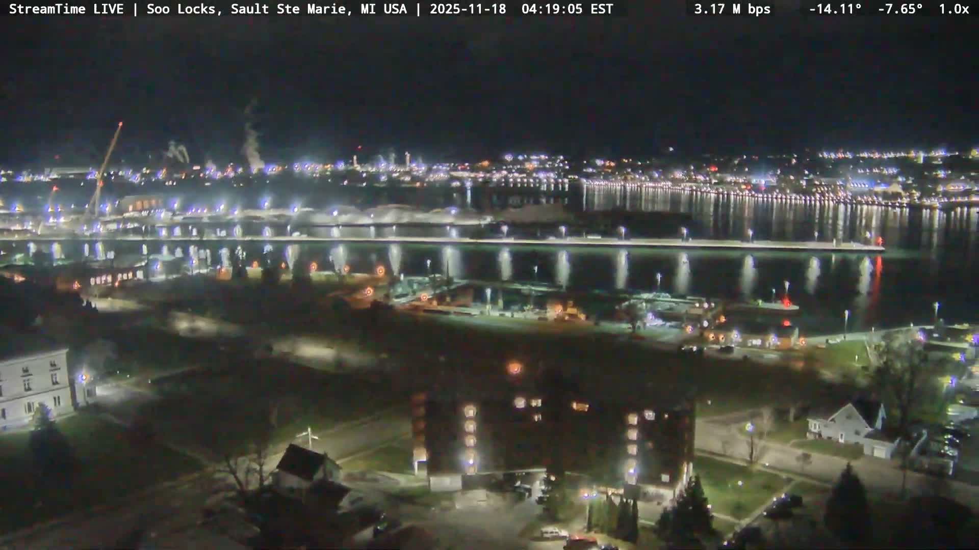 The image shows a clear, dark night view of a waterfront area with numerous lights illuminating buildings, docks with boats, and reflecting across the still water.