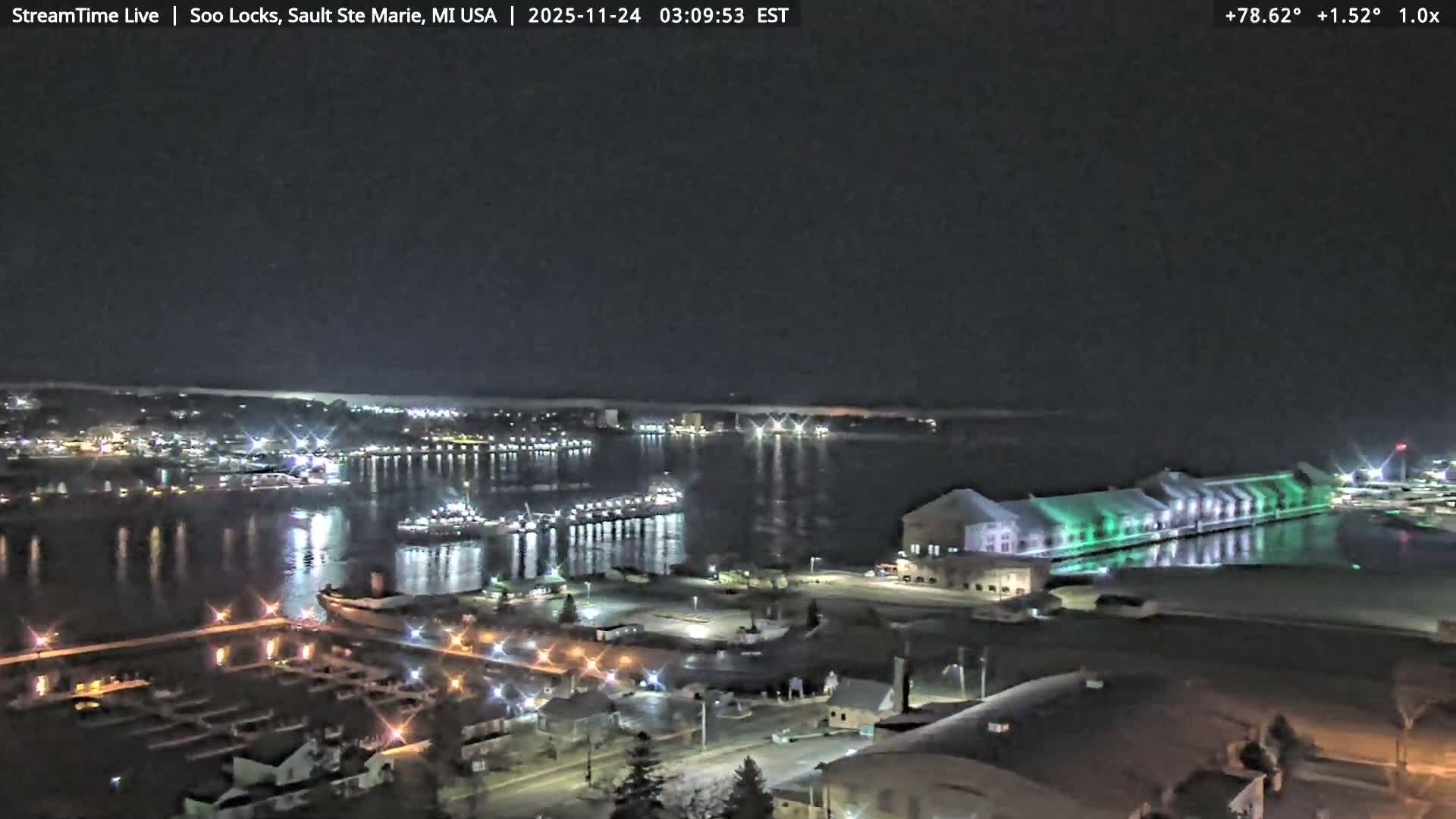 A clear night view captures a sprawling waterfront with city lights vividly reflecting on calm dark water, showcasing several docked ships and a long building illuminated in green.