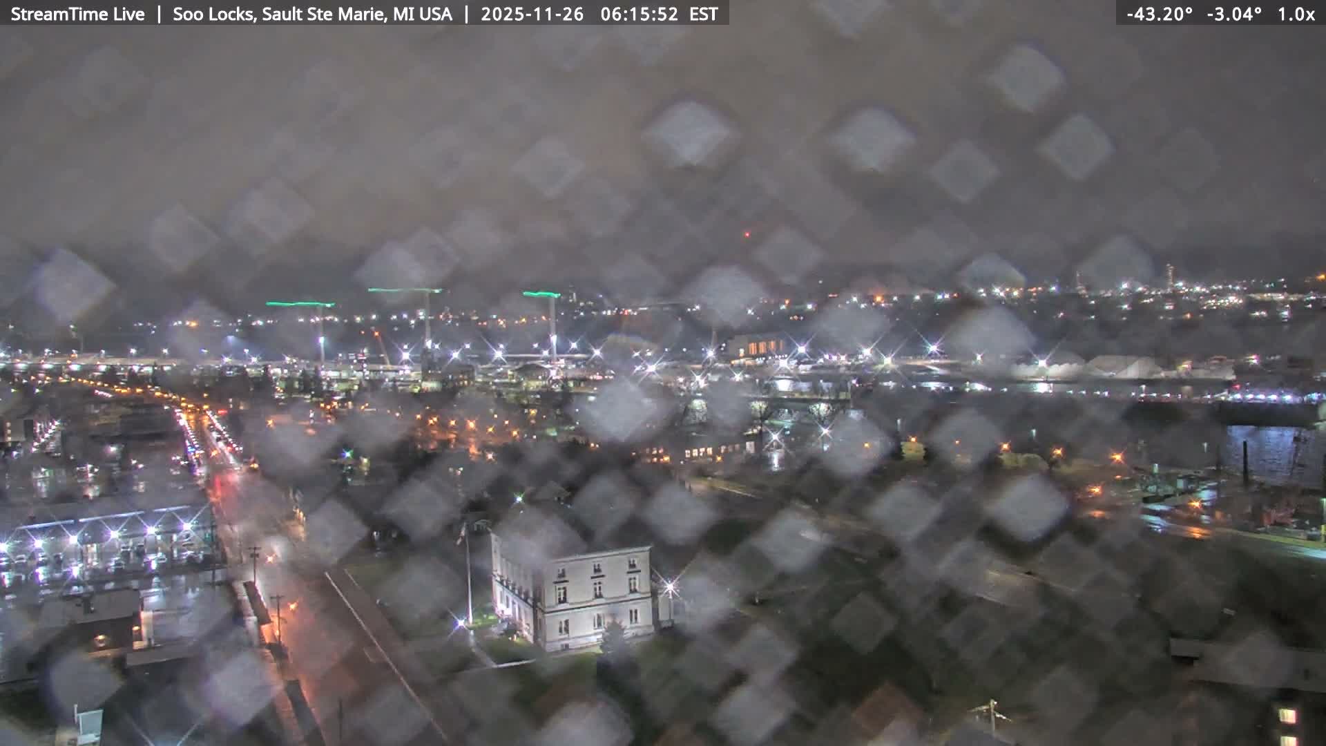 A clear night view captures a sprawling waterfront with city lights vividly reflecting on calm dark water, showcasing several docked ships and a long building illuminated in green.