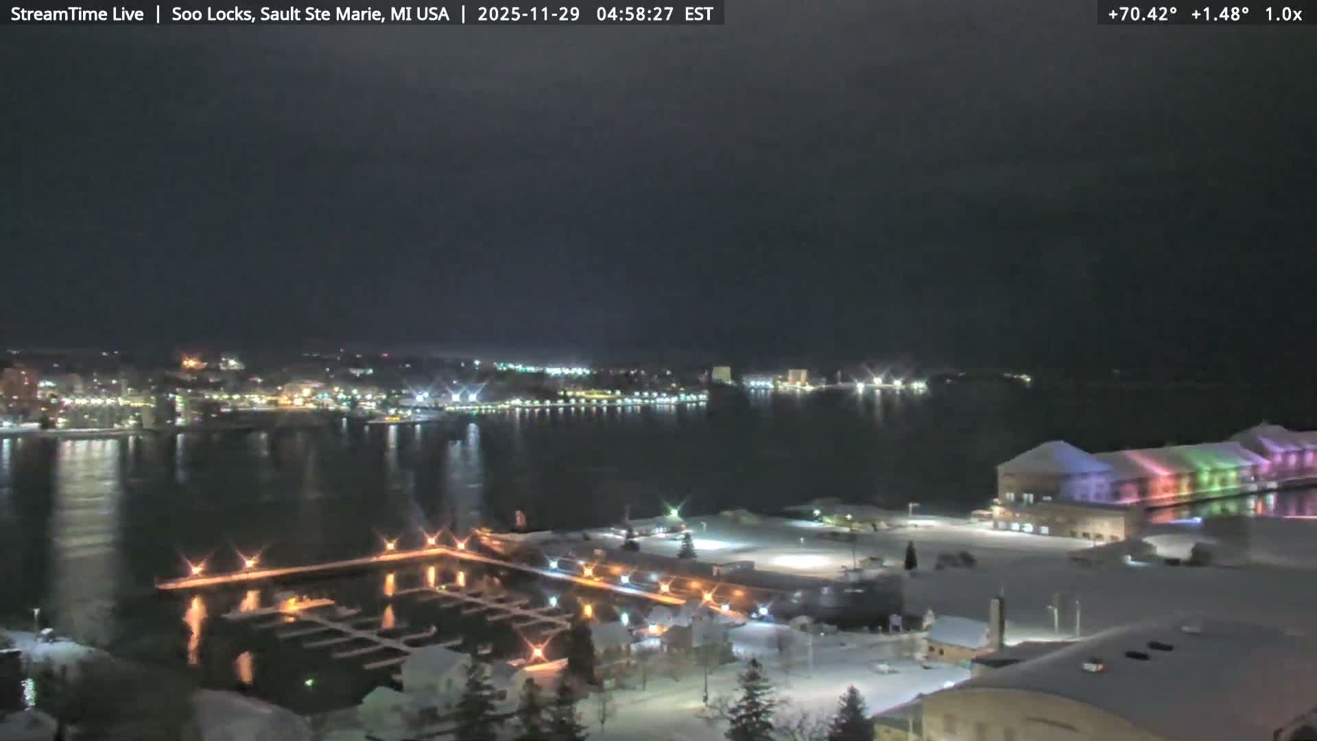 A snowy, cloudy winter night scene depicts a city skyline across dark water with numerous lights reflecting on its surface, a brightly lit marina, and a colorful illuminated building on the snow-covered near shore.