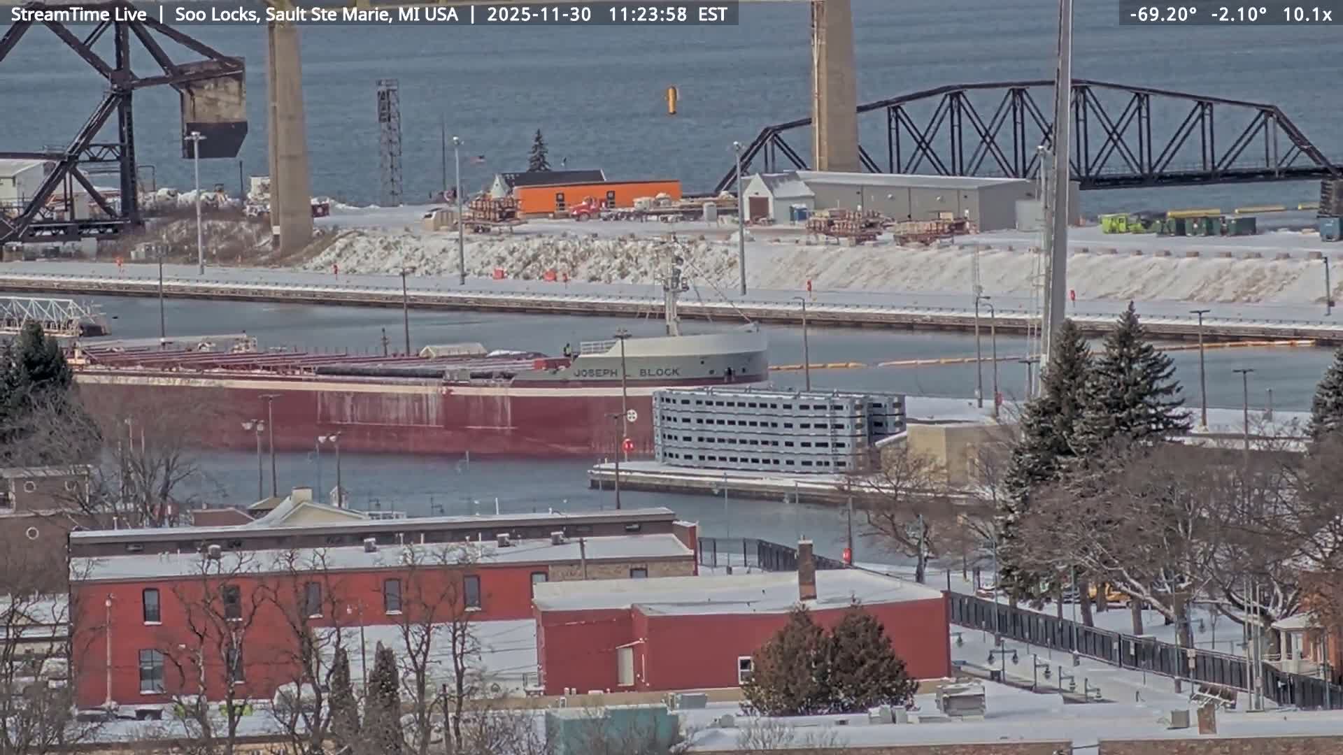 A snowy, cloudy winter night scene depicts a city skyline across dark water with numerous lights reflecting on its surface, a brightly lit marina, and a colorful illuminated building on the snow-covered near shore.