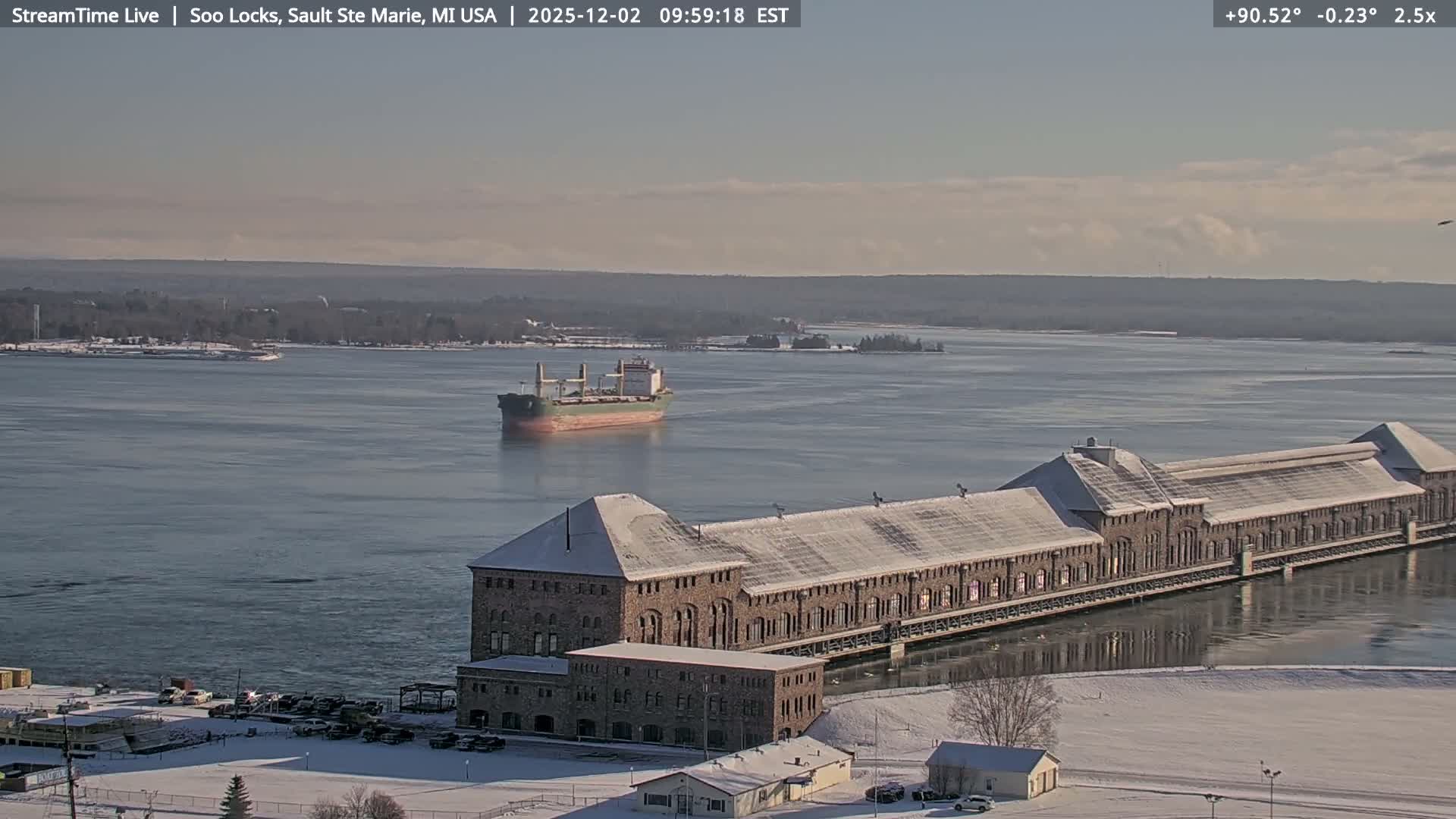 A cargo ship traverses a partially frozen waterway next to a long, snow-covered stone building, with the surrounding landscape dusted in snow under a bright, clear winter sky.