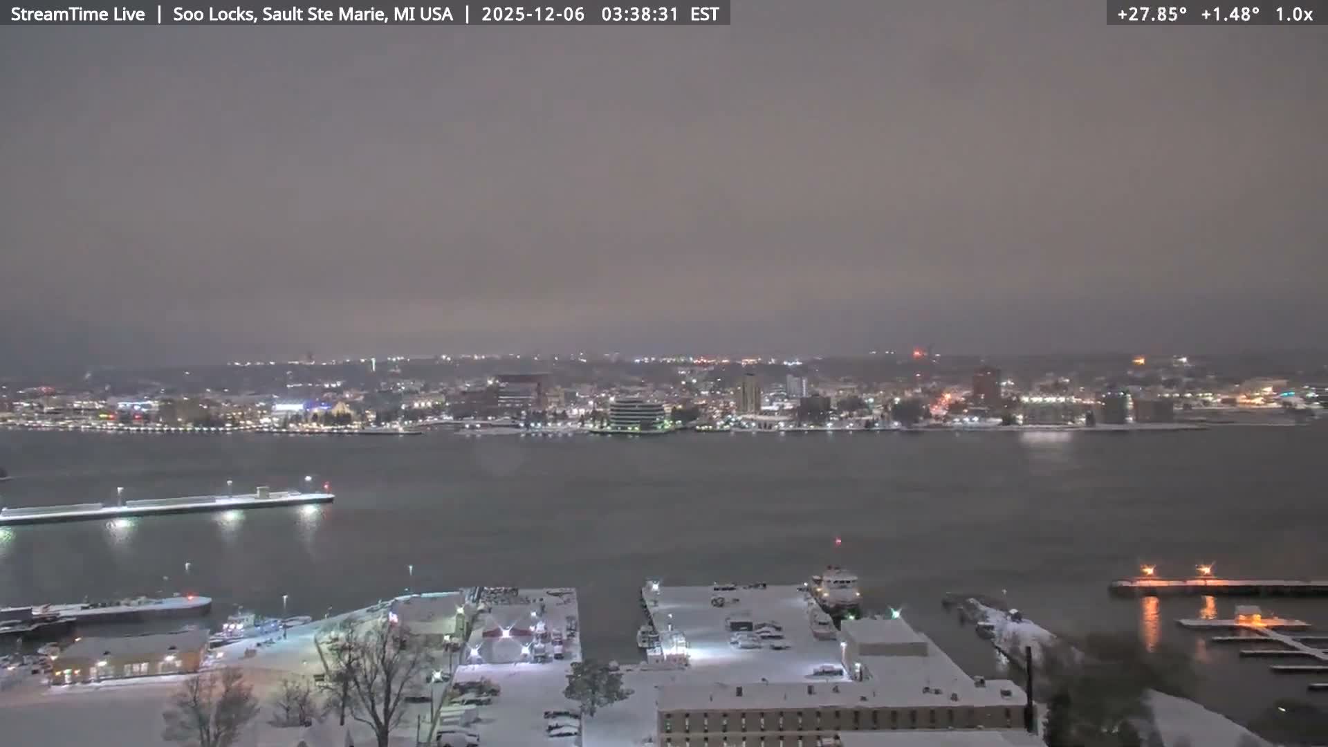 A cold, snowy night scene reveals a brightly lit city skyline across a dark body of water, featuring snow-covered docks and buildings in the foreground under an overcast sky.