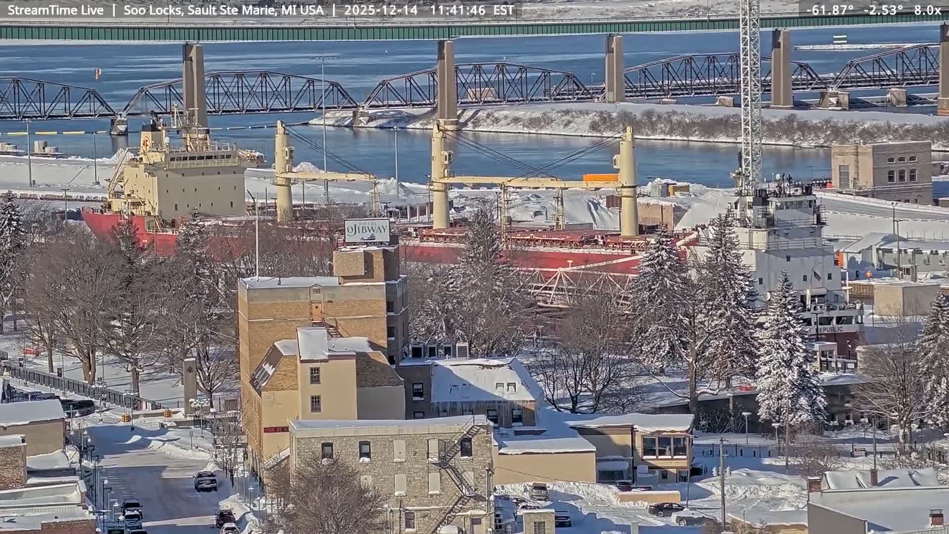 A dark, snowy, and very foggy outdoor scene at night shows numerous artificial lights reflecting on a body of water, with faint shapes of buildings and trees barely visible through the thick atmosphere.