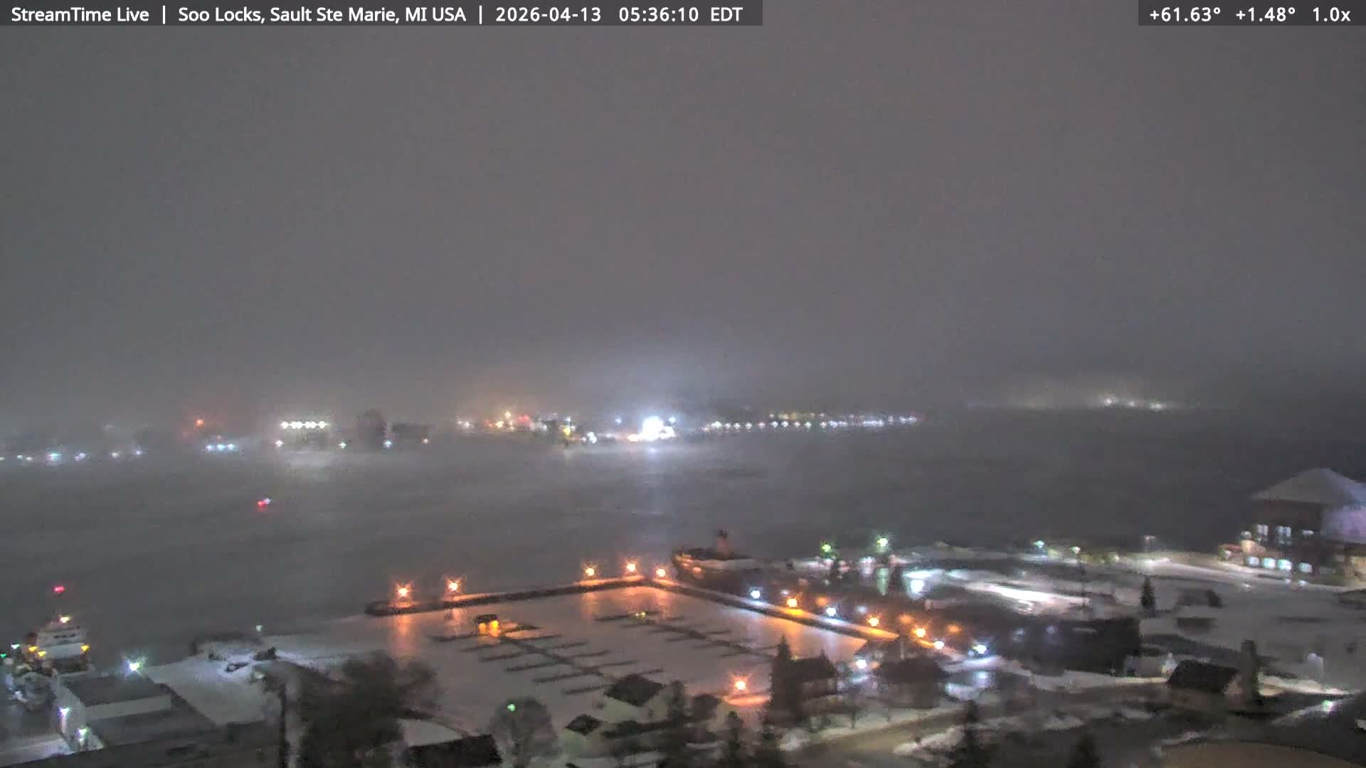 A dark, snowy, and very foggy outdoor scene at night shows numerous artificial lights reflecting on a body of water, with faint shapes of buildings and trees barely visible through the thick atmosphere.