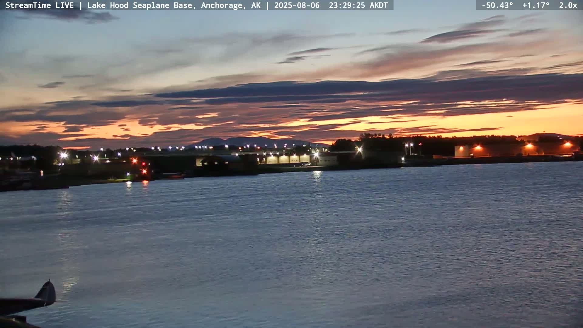 A twilight scene shows a body of water with a building complex and distant mountains under a partly cloudy sky.