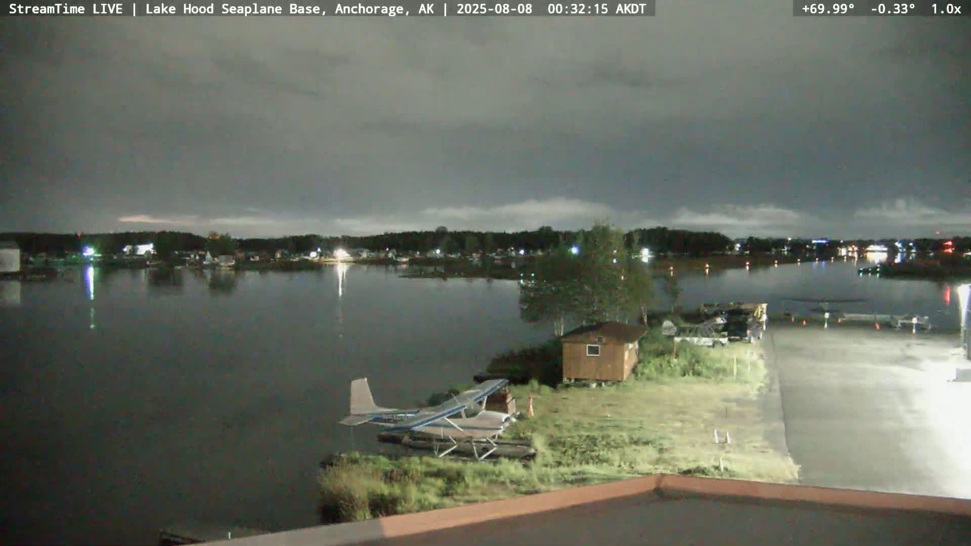 A seaplane is parked near a small building next to a lake at night under a cloudy sky.