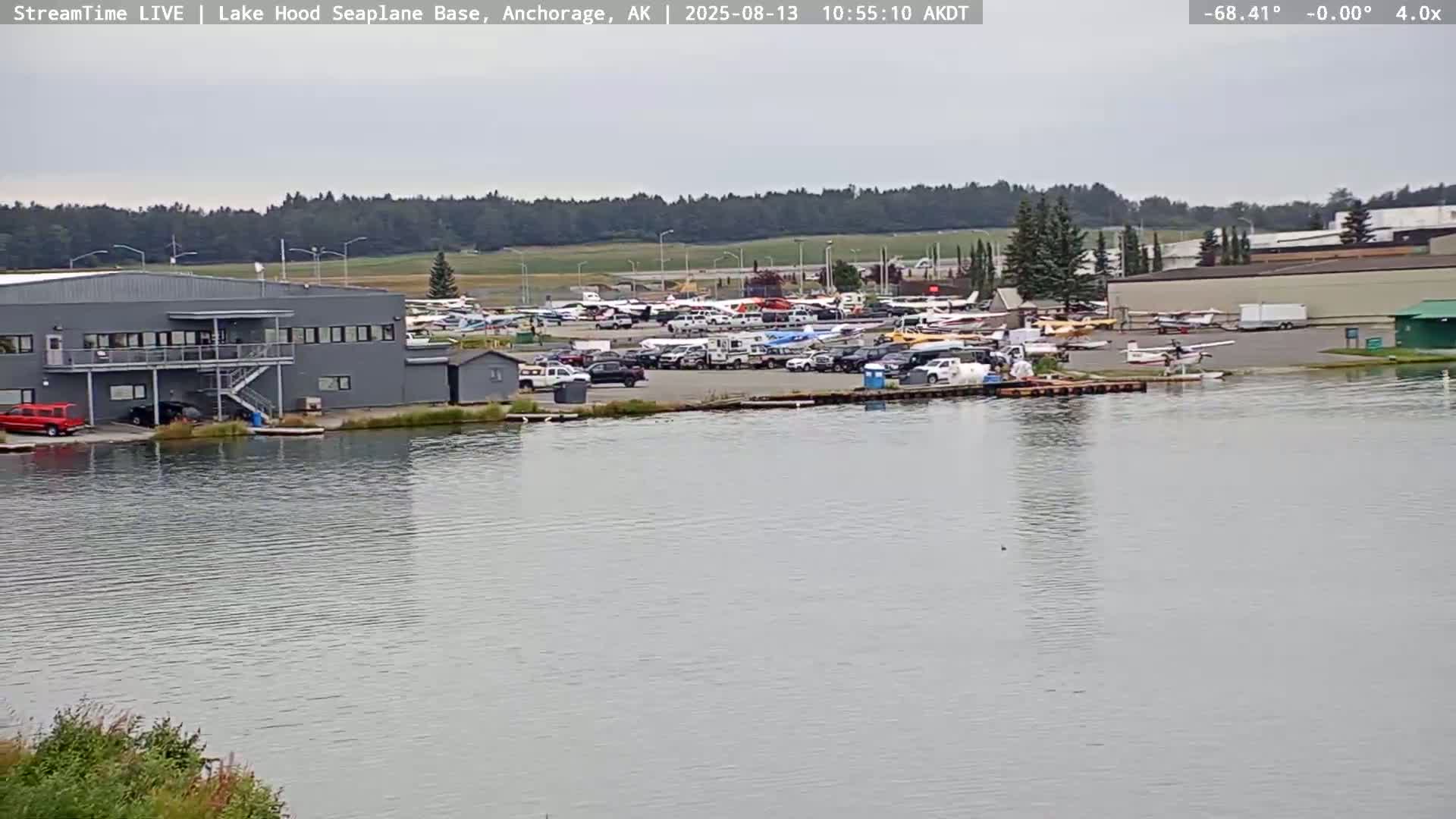 A seaplane base with numerous aircraft and vehicles is seen on a cloudy day next to a calm body of water.