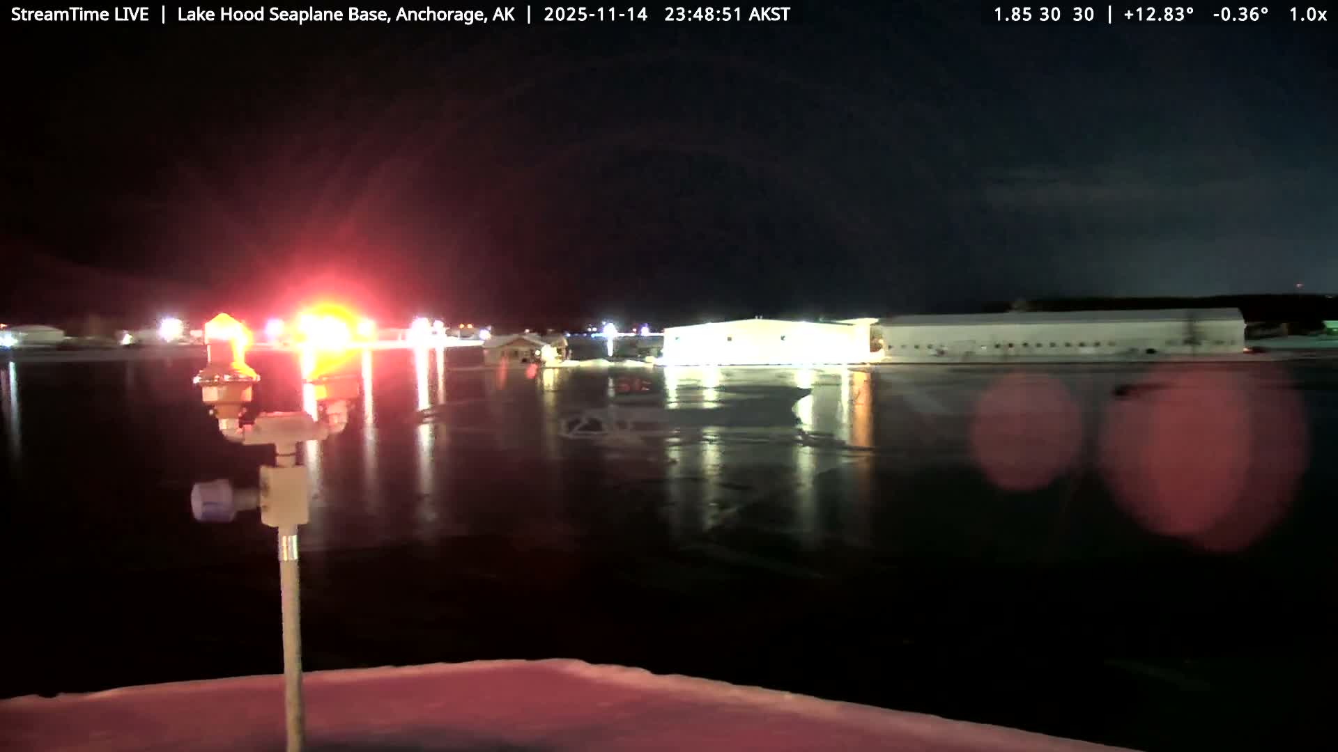 A clear, cold night at Lake Hood Seaplane Base reveals bright red and orange airfield lights reflecting on a dark, partially frozen surface with snow in the foreground, and illuminated buildings in the distance under a dark sky.