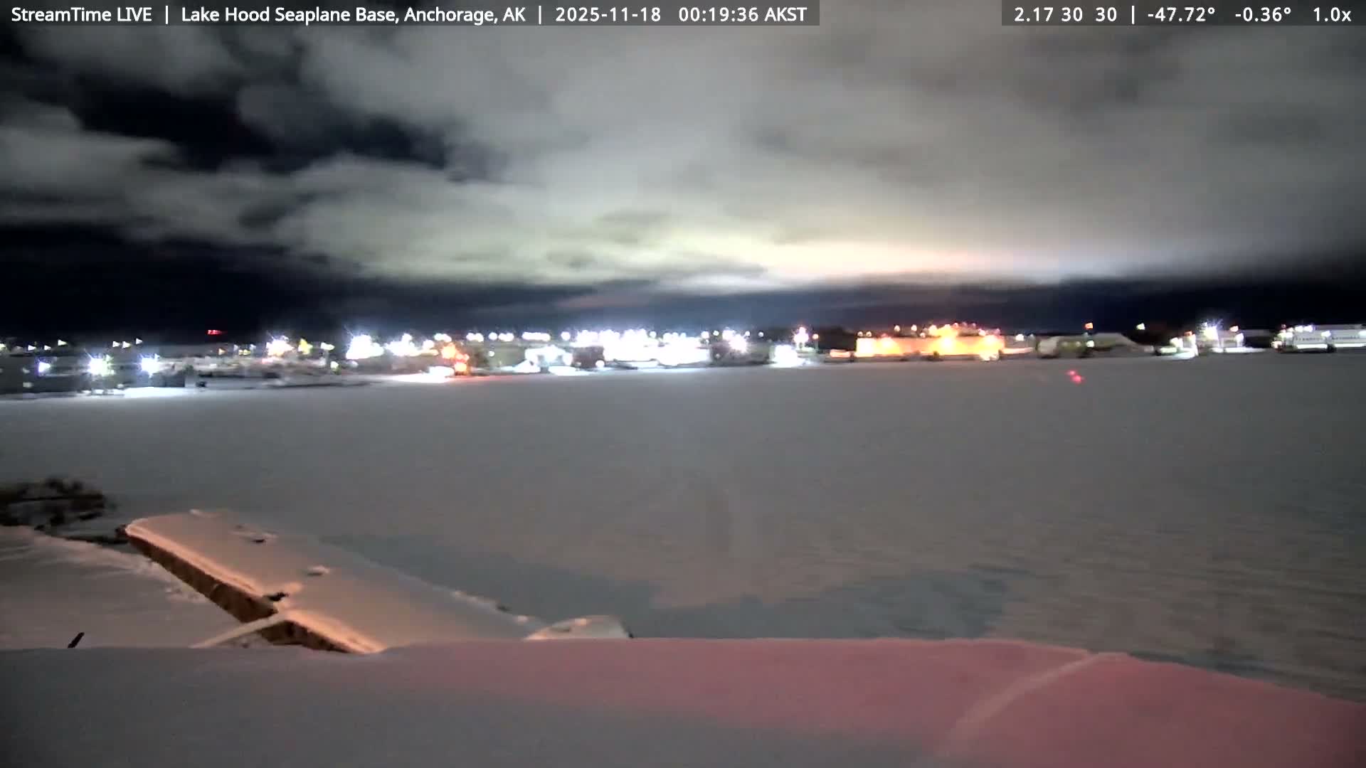 A clear, cold night at Lake Hood Seaplane Base reveals bright red and orange airfield lights reflecting on a dark, partially frozen surface with snow in the foreground, and illuminated buildings in the distance under a dark sky.