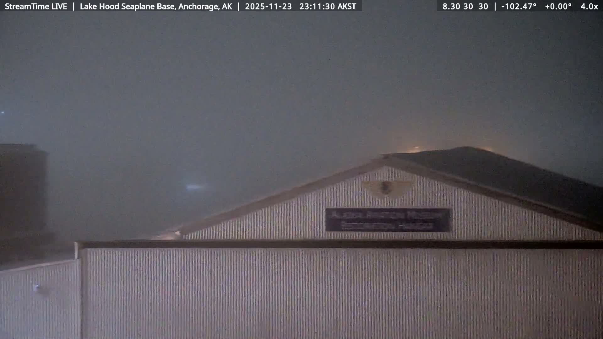 A corrugated metal building with a dark sloped roof is partially visible at night through a dense fog, with faint lights glowing through the hazy conditions.