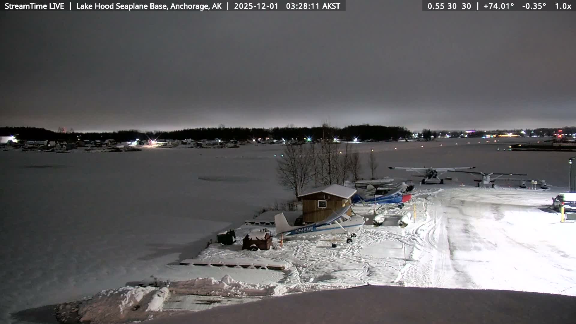 The image displays a dark, snowy night at a seaplane base with several small planes parked on a snow-covered ramp next to a frozen lake, while distant town lights twinkle under an overcast sky.