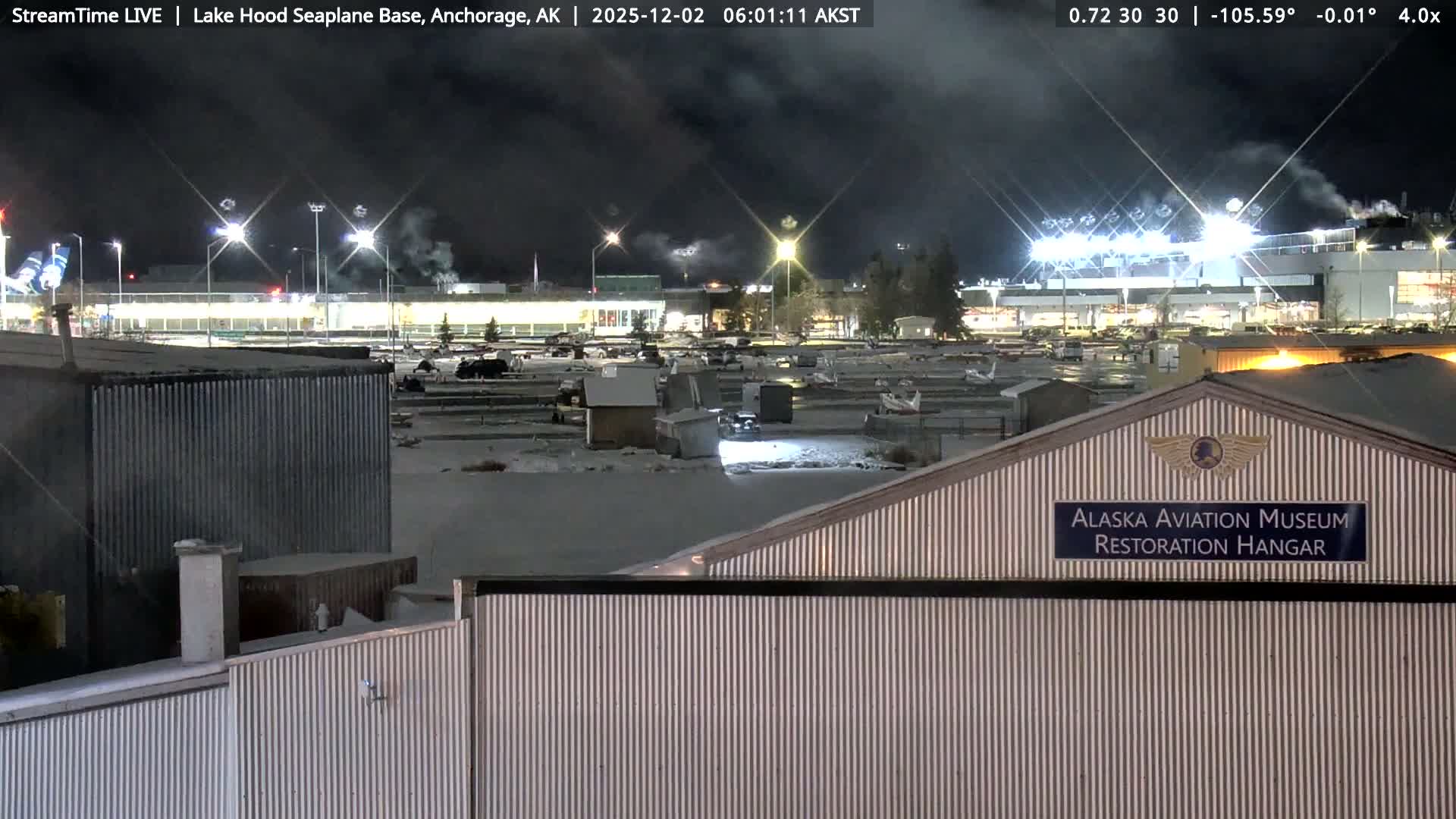A nighttime image shows an illuminated seaplane base with snow and ice covering the ground and parked aircraft, featuring a corrugated metal aviation museum hangar in the foreground and steam rising from distant industrial-looking structures under a dark, cloudy sky.