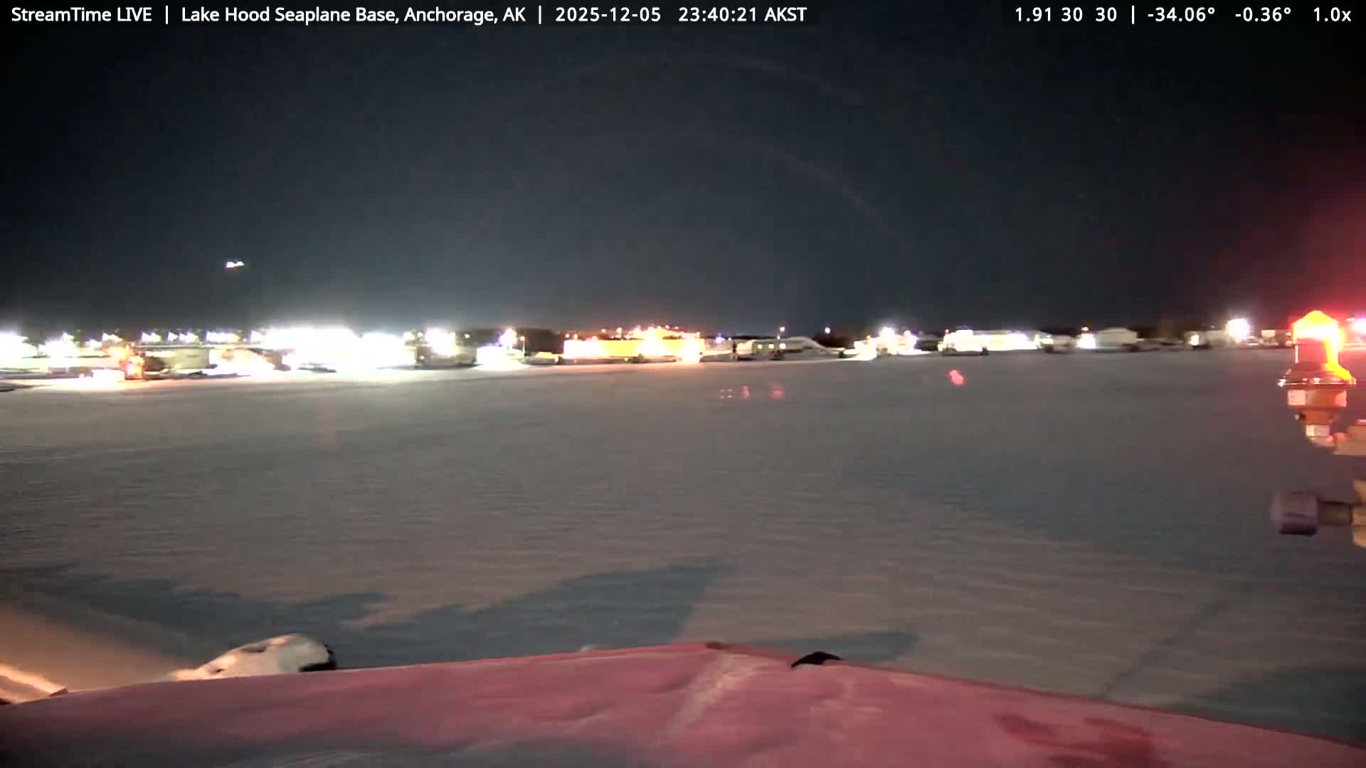 The image captures a vast, snow-covered airfield at night under a clear, dark sky, with numerous bright lights illuminating distant structures and a red beacon visible in the foreground, indicating clear and cold winter weather.