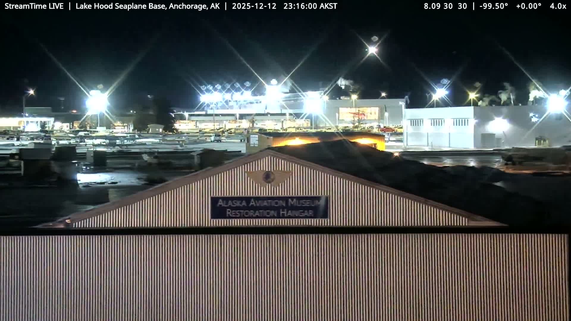 A clear night illuminates a striped building in the foreground, with numerous brightly lit structures and a parking area visible in the background at what appears to be an industrial or airfield complex.