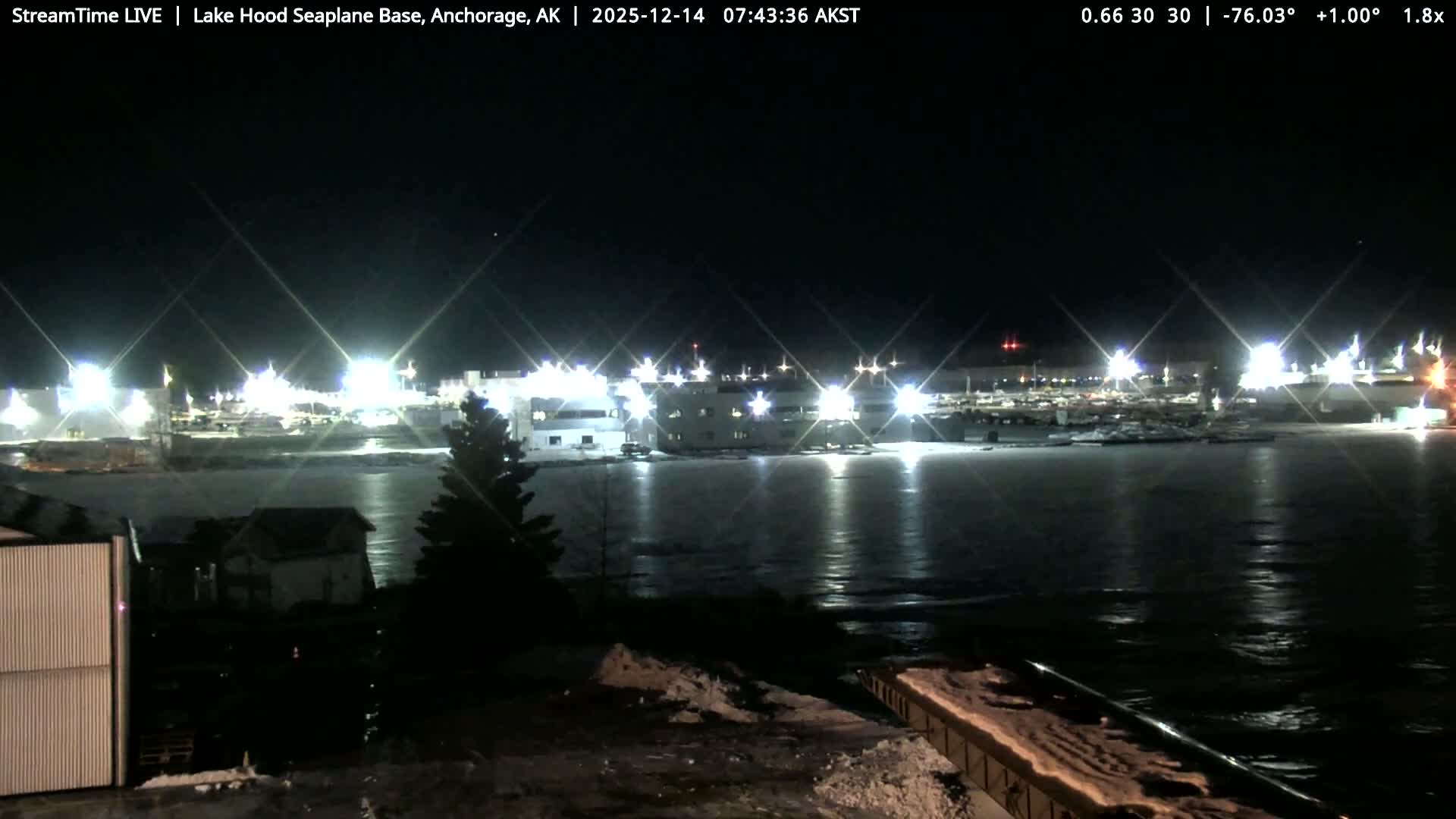 A clear night illuminates a striped building in the foreground, with numerous brightly lit structures and a parking area visible in the background at what appears to be an industrial or airfield complex.